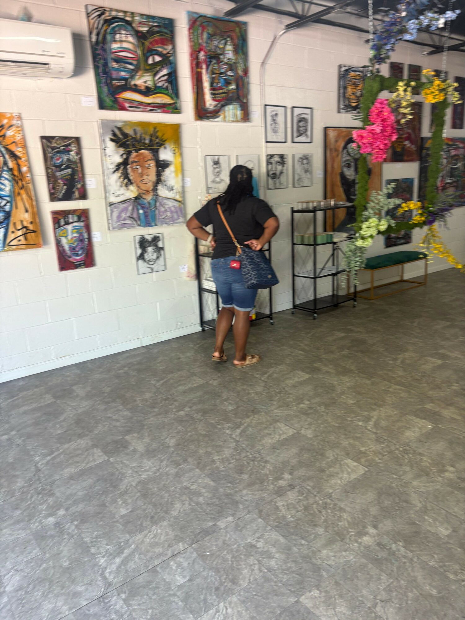 Person standing in an art gallery, looking at paintings on the wall, with flowers on a table nearby.