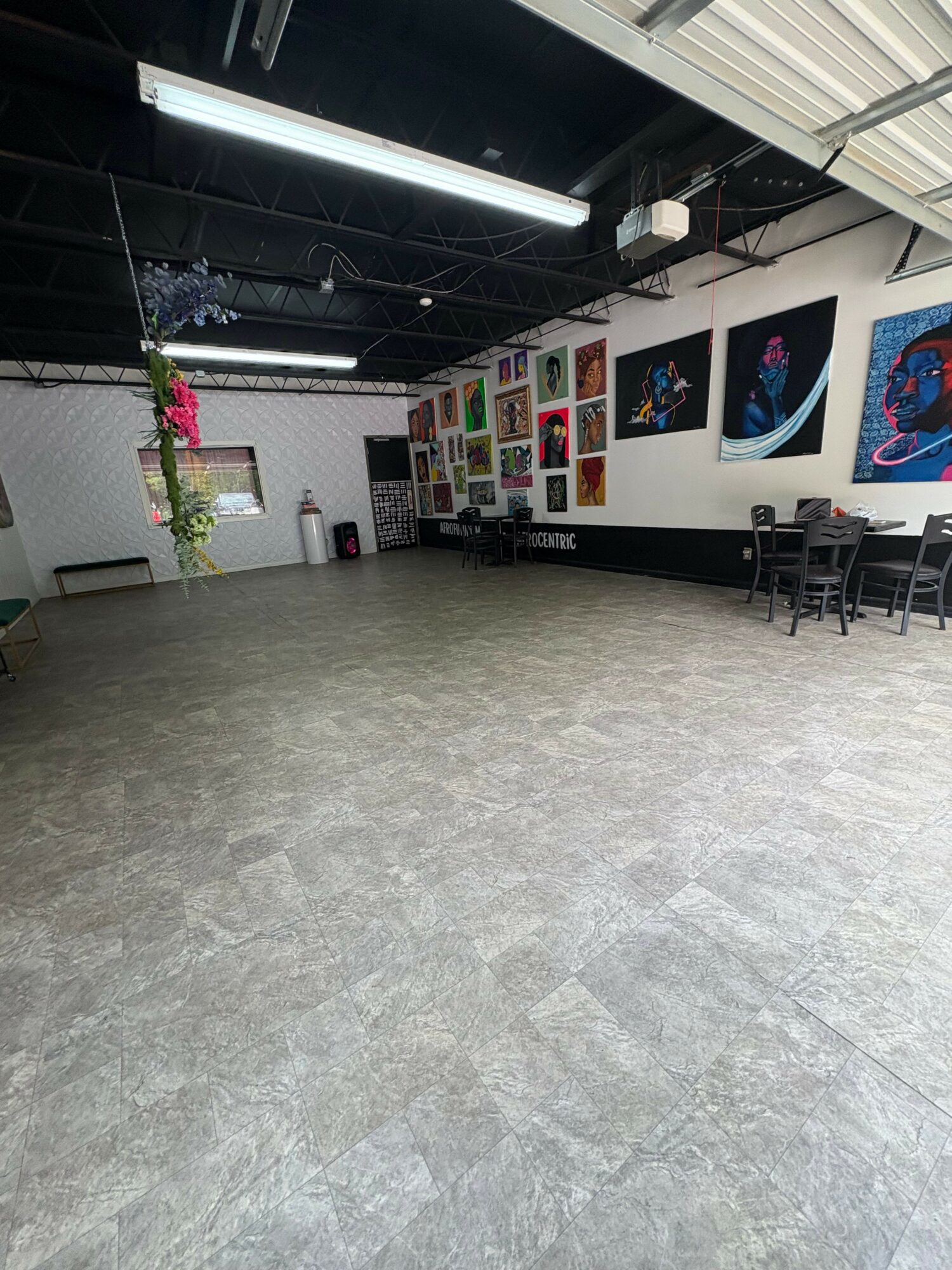 Art gallery interior with colorful paintings on white walls and a gray floor, ceiling with black beams and lighting.