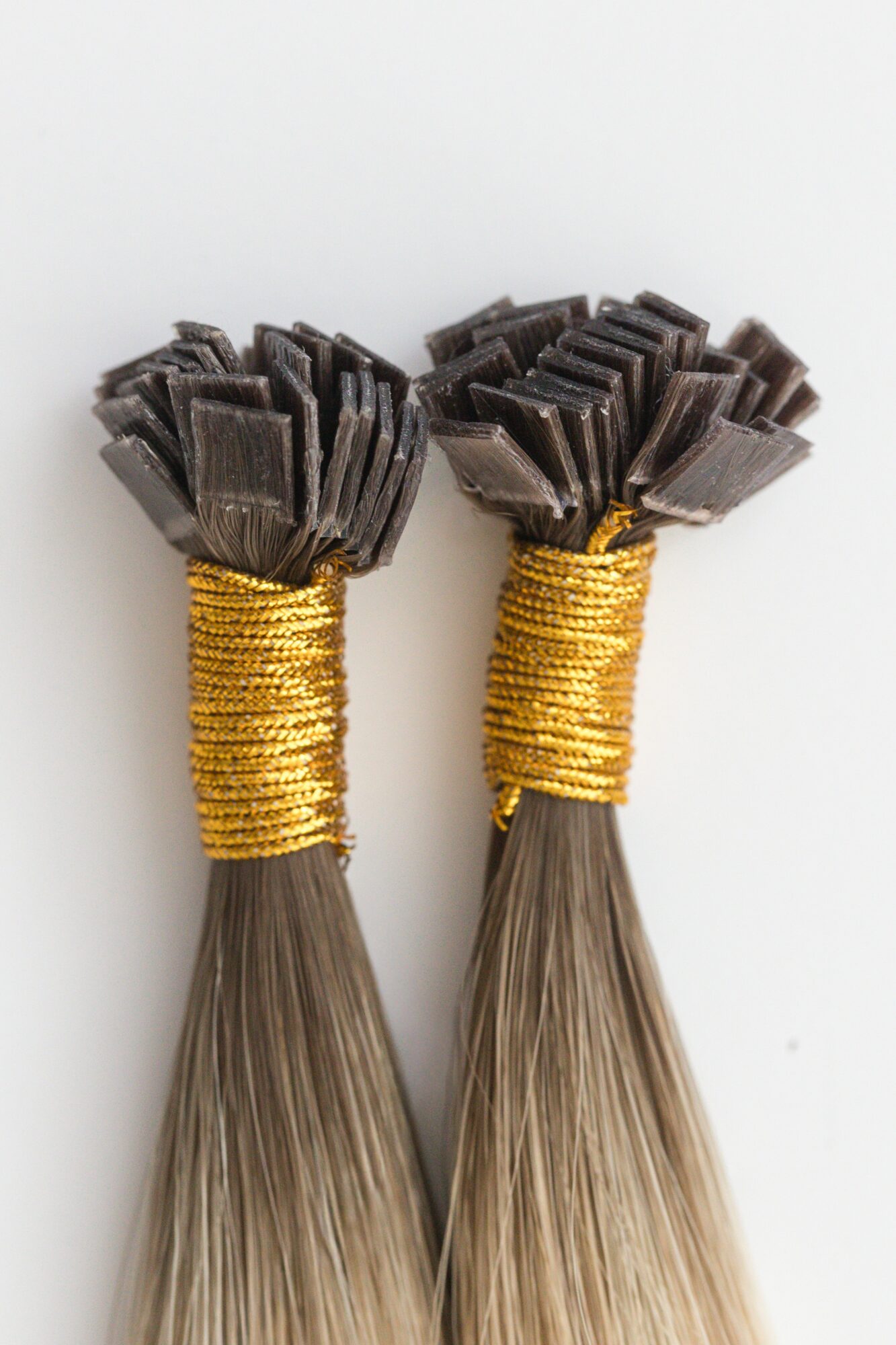 Two hair extensions with dark brown tips, gold thread wrapping around the top, and long straight hair strands.