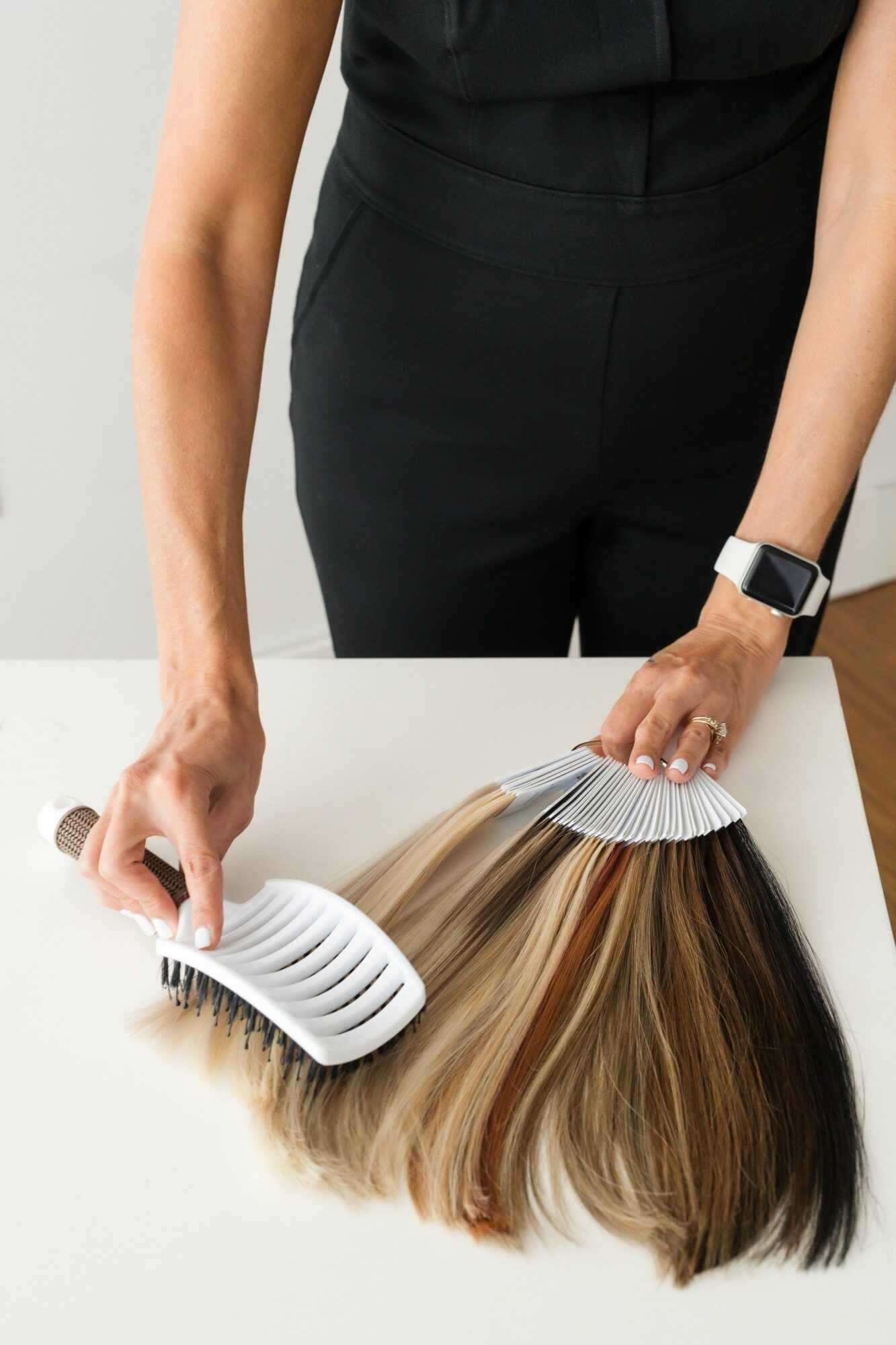 Person with black clothing and smartwatch holds a hair color sample fan and a hairbrush on a white surface.
