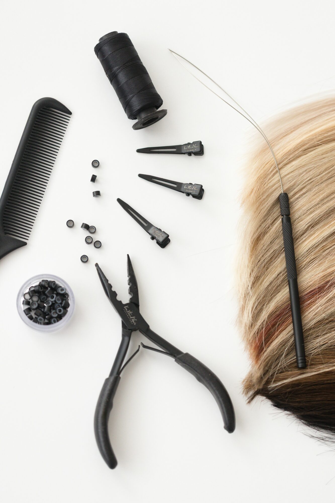 Hairdresser's tools including a comb, scissors, a spool of thread, and a section of blonde hair with a hairbrush, on a white background.