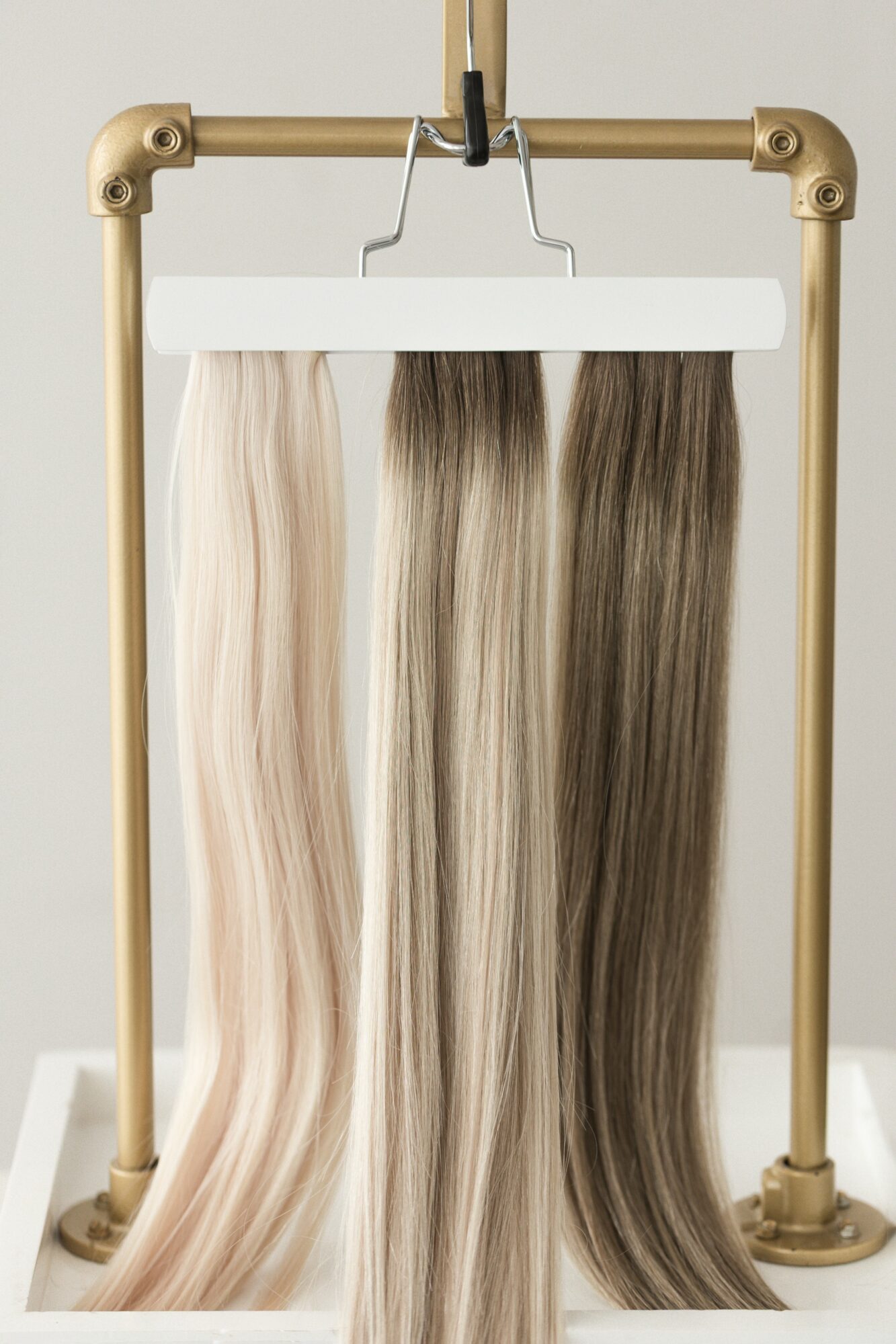 Four hair color samples hanging on a white hanger, from light blonde to dark brown, on a gold-colored stand.