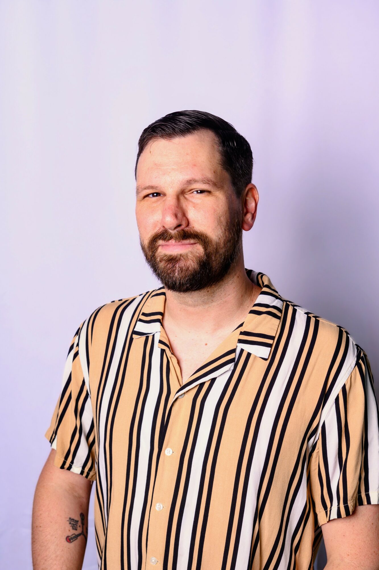 Man with dark hair and beard wearing a striped shirt, smiling, against a light background.