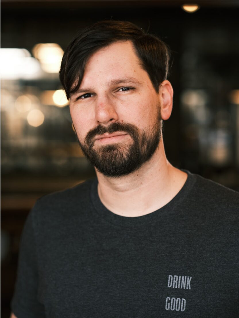 Man with dark hair and beard wearing a black shirt with text, standing indoors with blurred background.