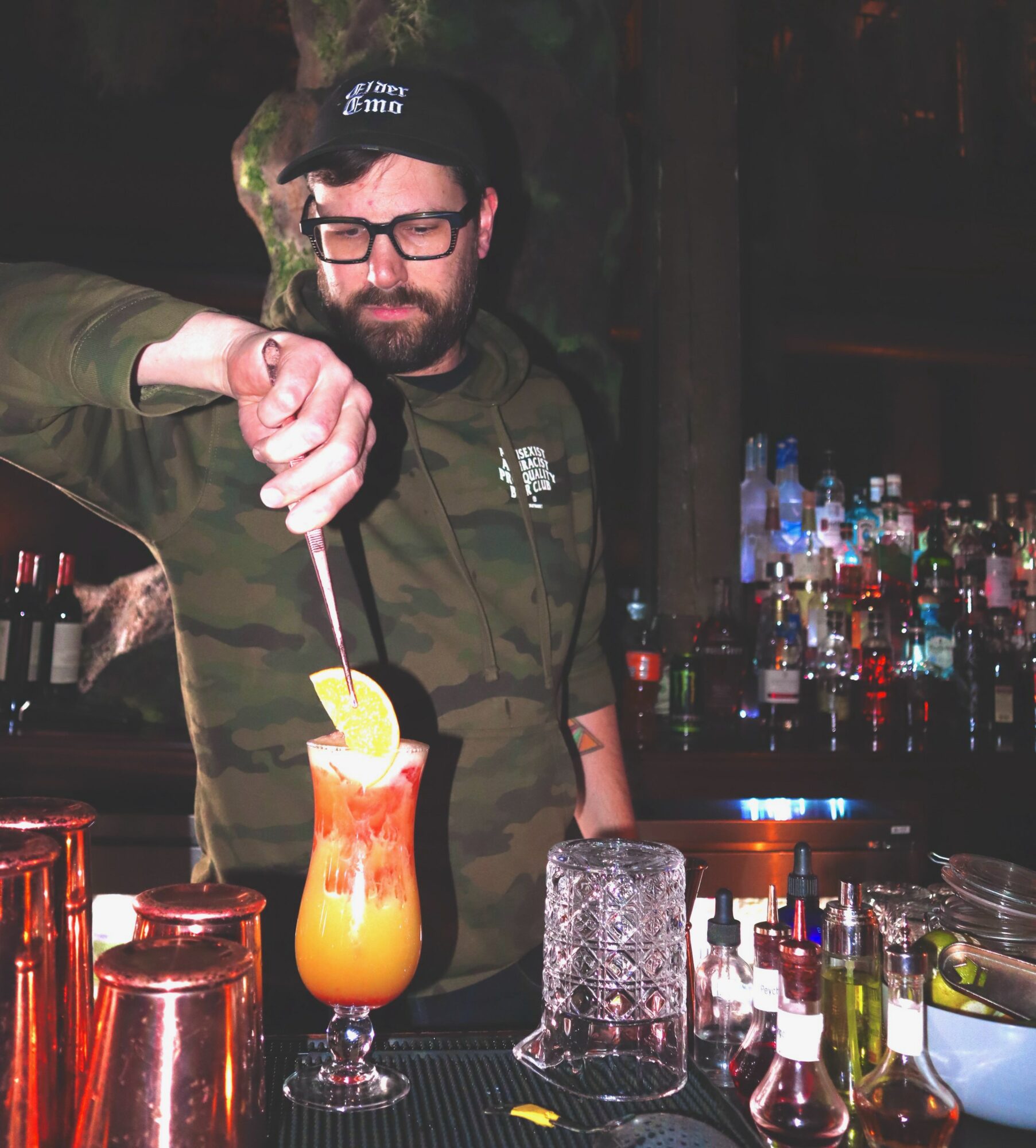 Man with glasses and a beard prepares a colorful cocktail at a bar, with bottles and bar tools in the background.