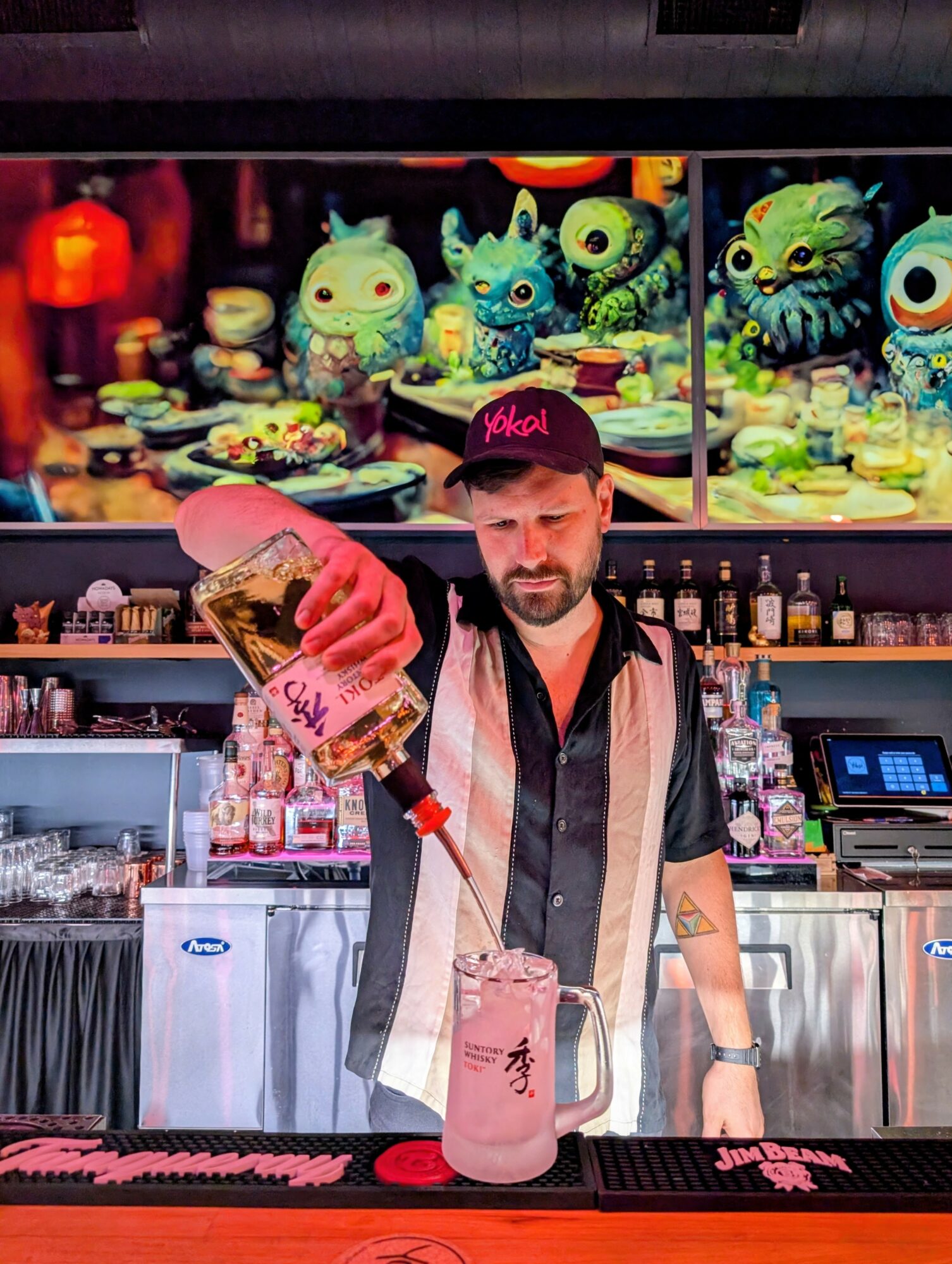 Bartender pouring drink at bar with colorful animated characters on screen behind him.