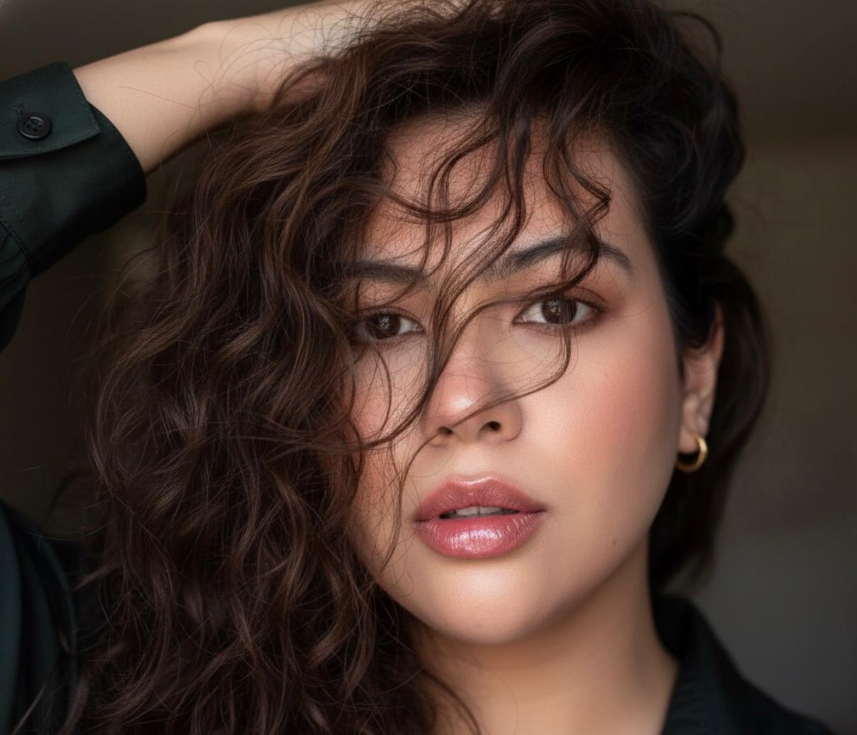 Close-up portrait of a woman with curly hair, looking directly at the camera, with a neutral background.