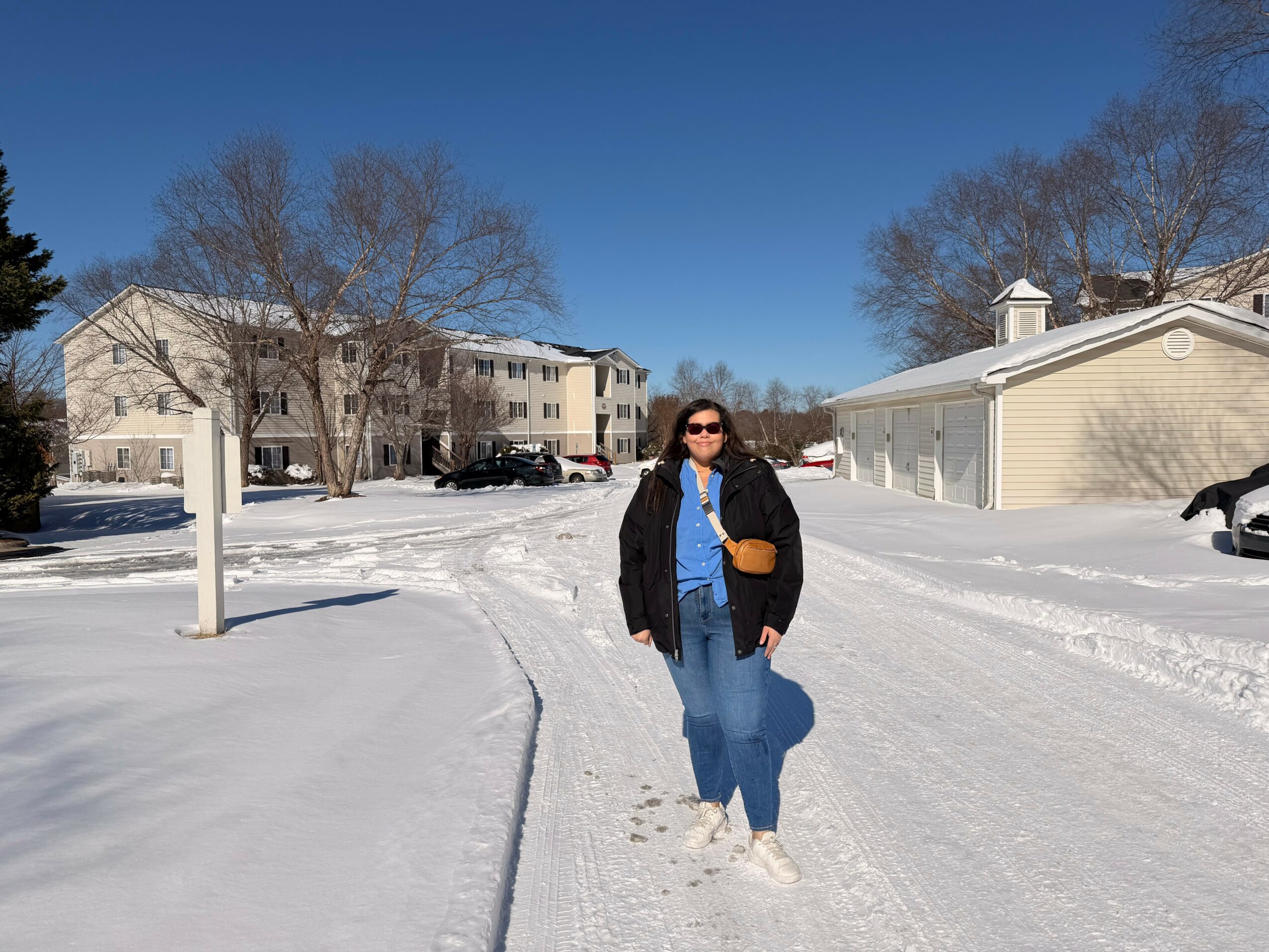 Person wearing sunglasses, black jacket, and blue jeans walking on snowy street with houses and trees in background.