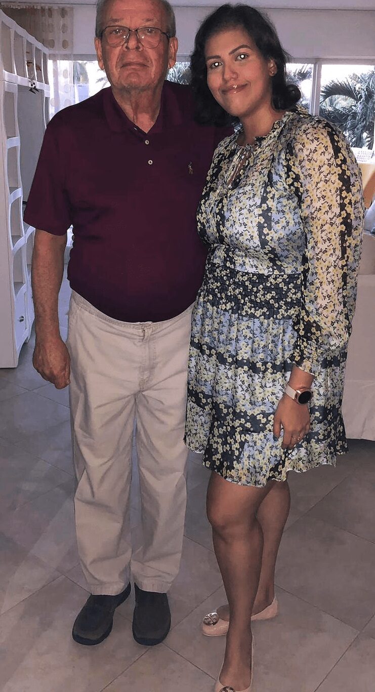 Older man and young woman standing indoors, smiling, with a window and furniture in the background.