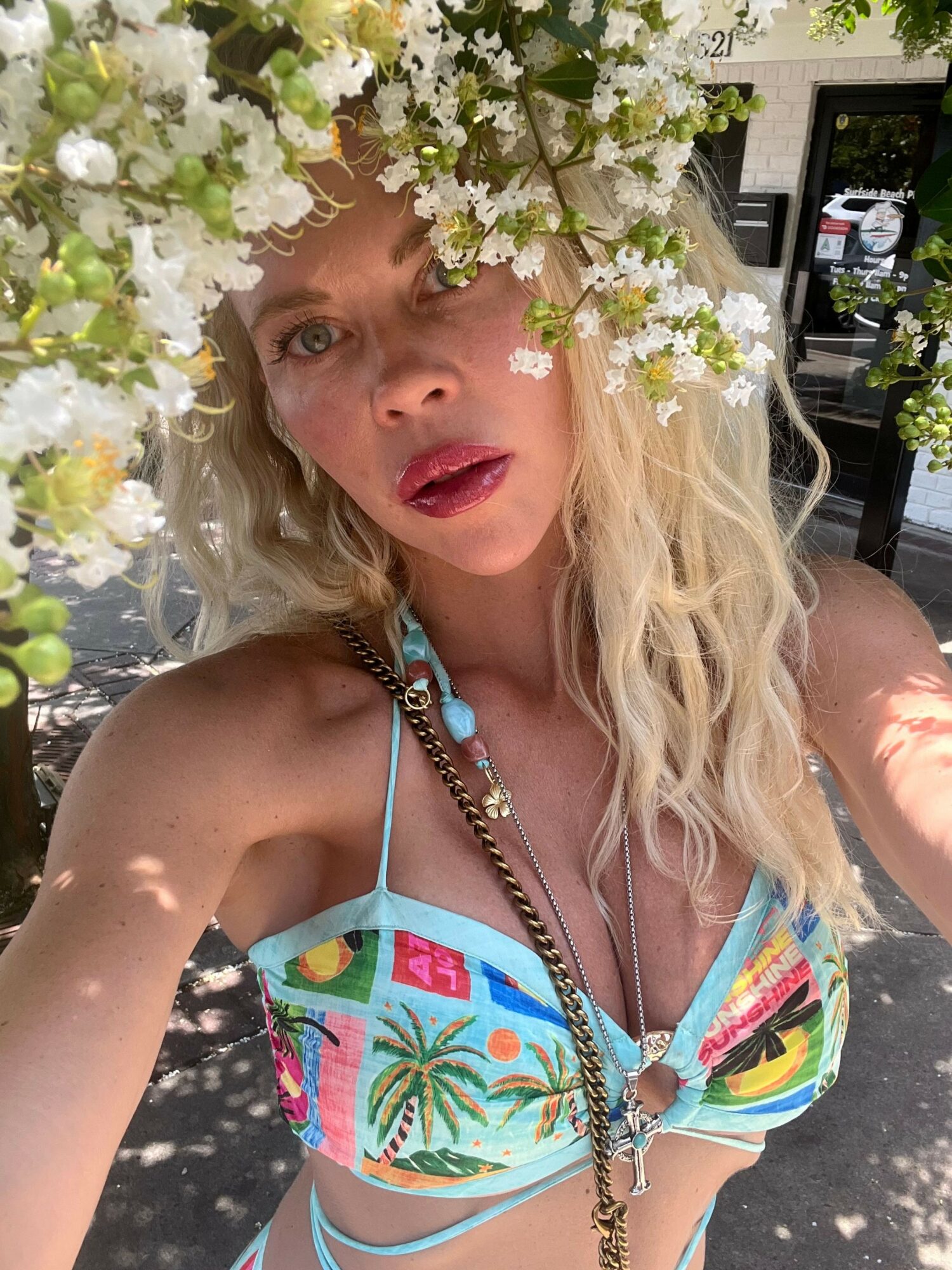 Woman with long blonde hair lying among white flowers, wearing a colorful bikini top, looking at the camera.