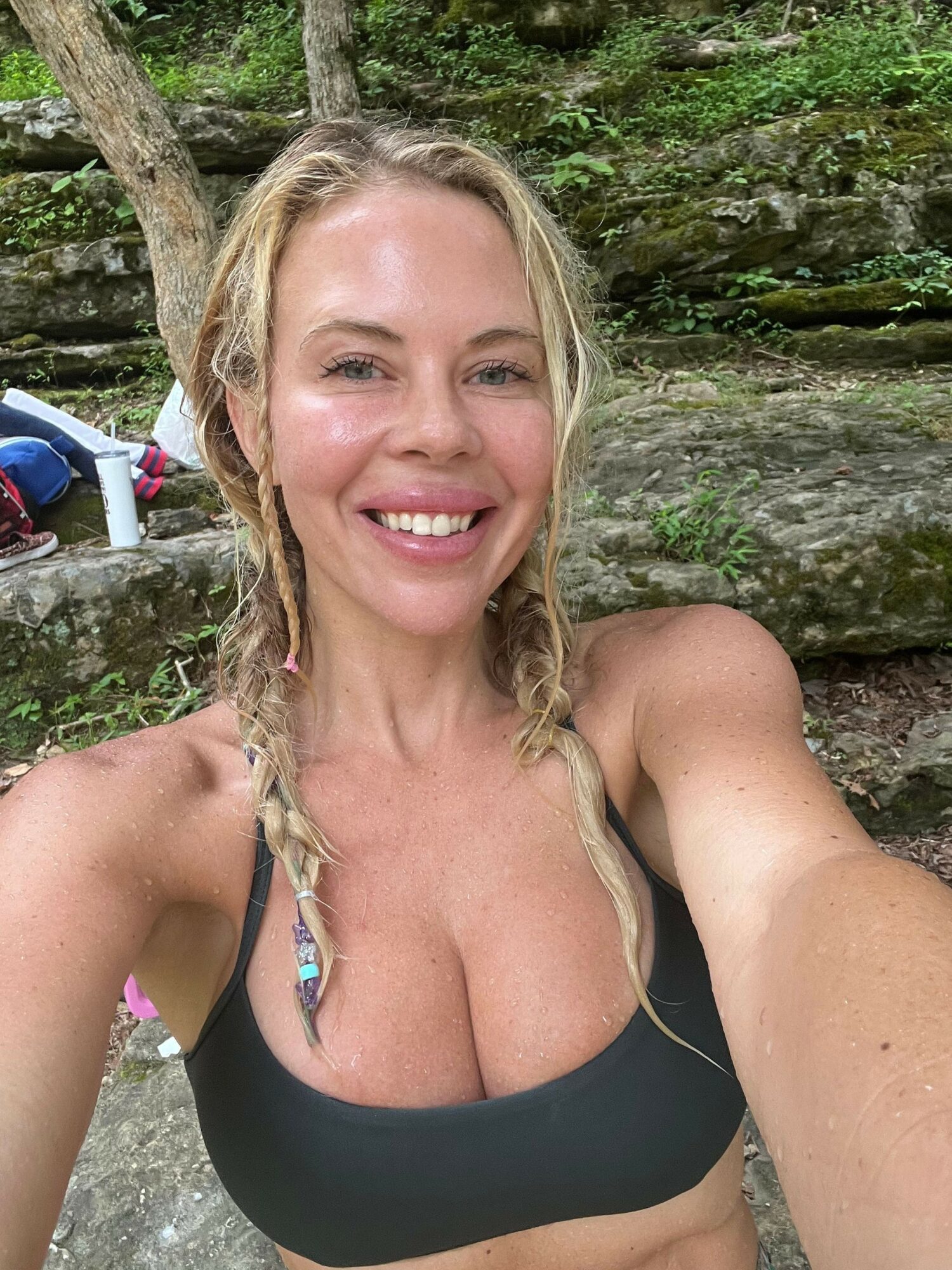 Woman with blonde hair in a braid smiling outdoors, wearing a black top, near a mossy rock wall.