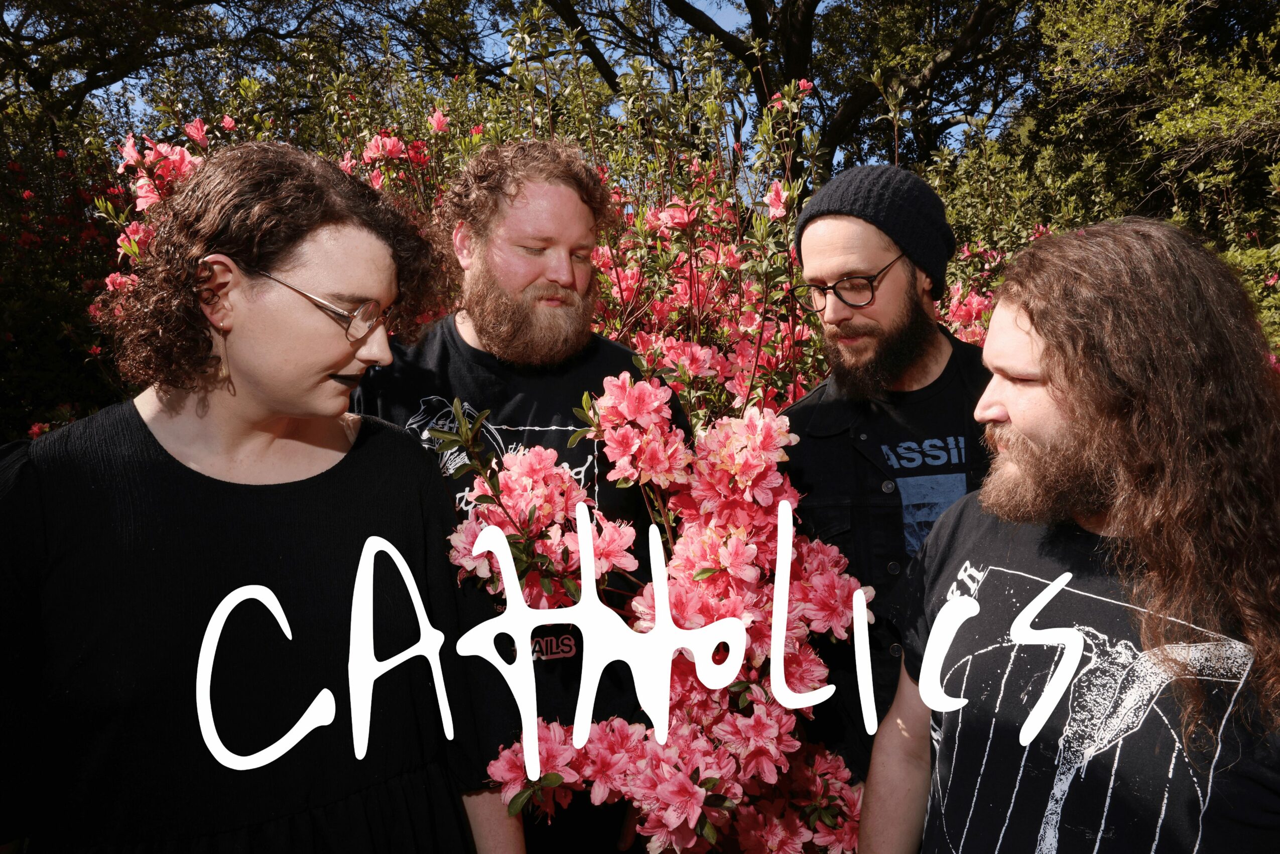 Four people with beards and glasses gather around pink flowers outdoors, engaging in conversation, with trees in the background.