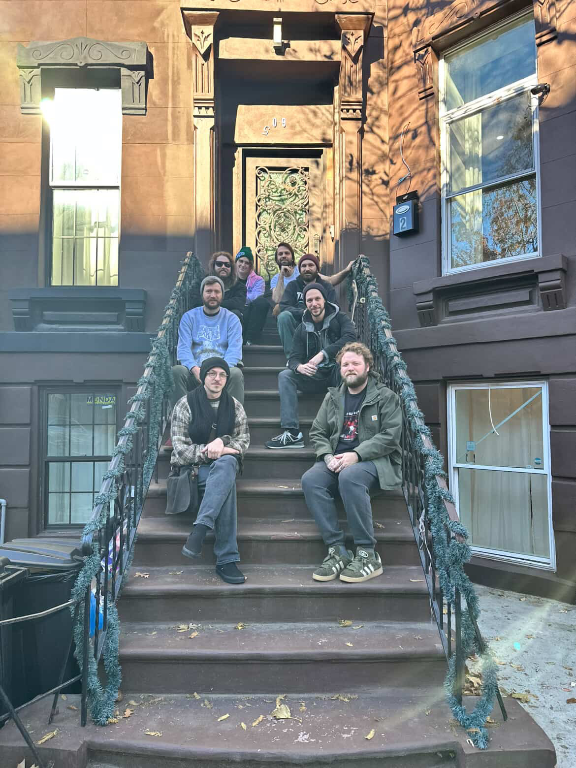 Group of seven people sitting on outdoor stairs in front of a brownstone building, some smiling, with windows and a door behind them.