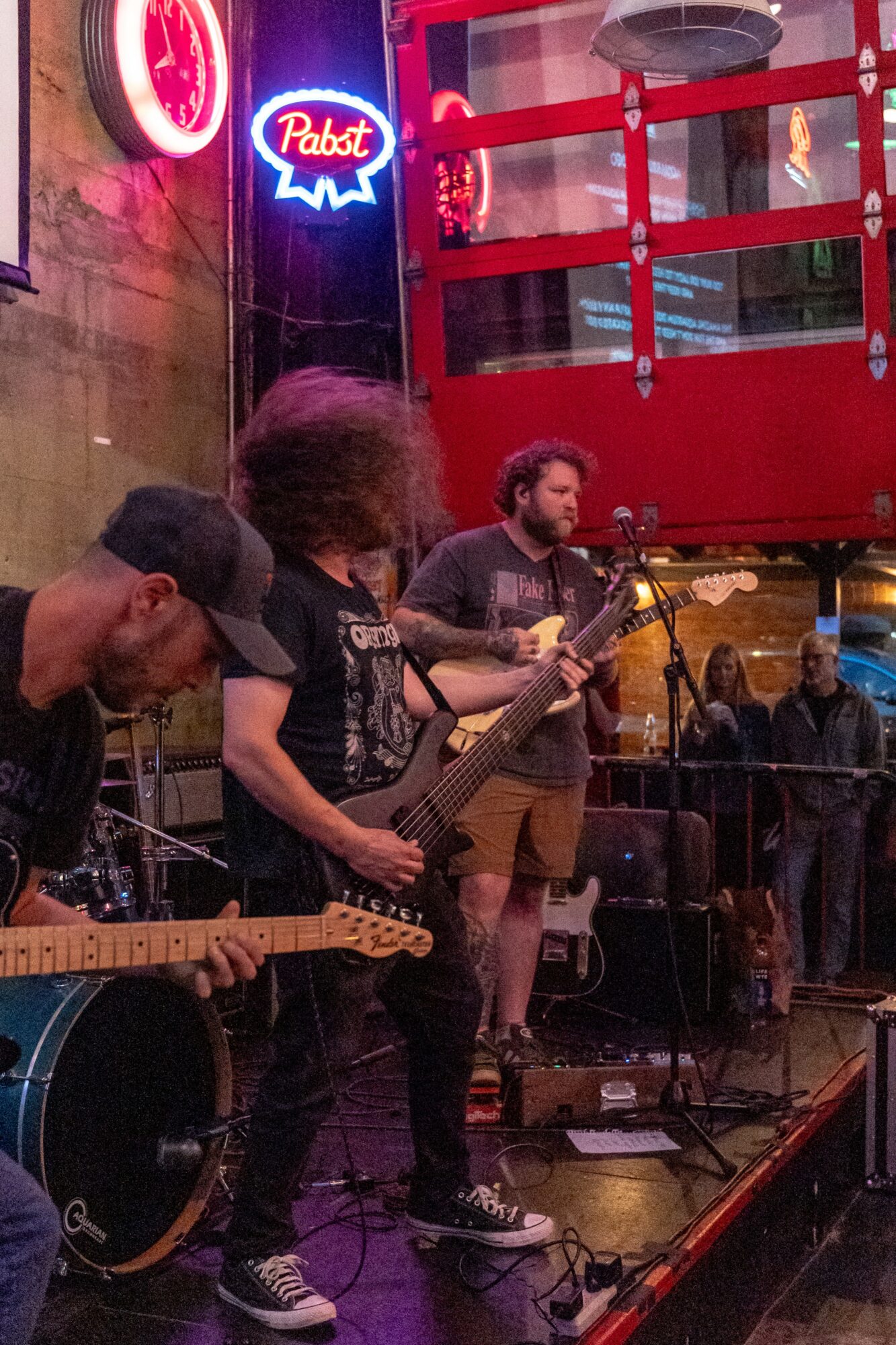 Musicians perform live with guitars in a dimly lit venue, neon signs and a red structure in the background.