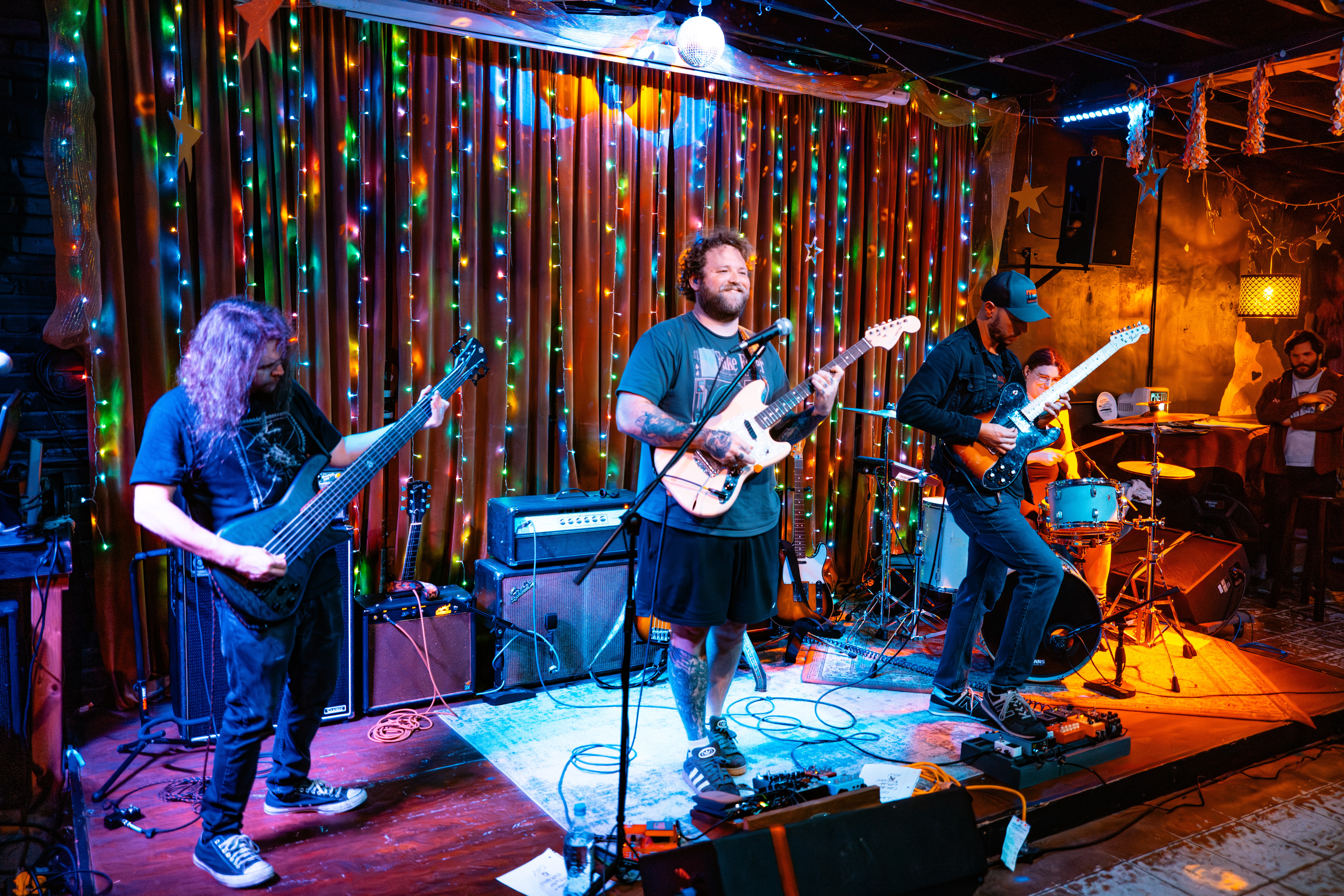 Four musicians perform on stage with colorful lights and a curtain backdrop, playing guitars and drums.