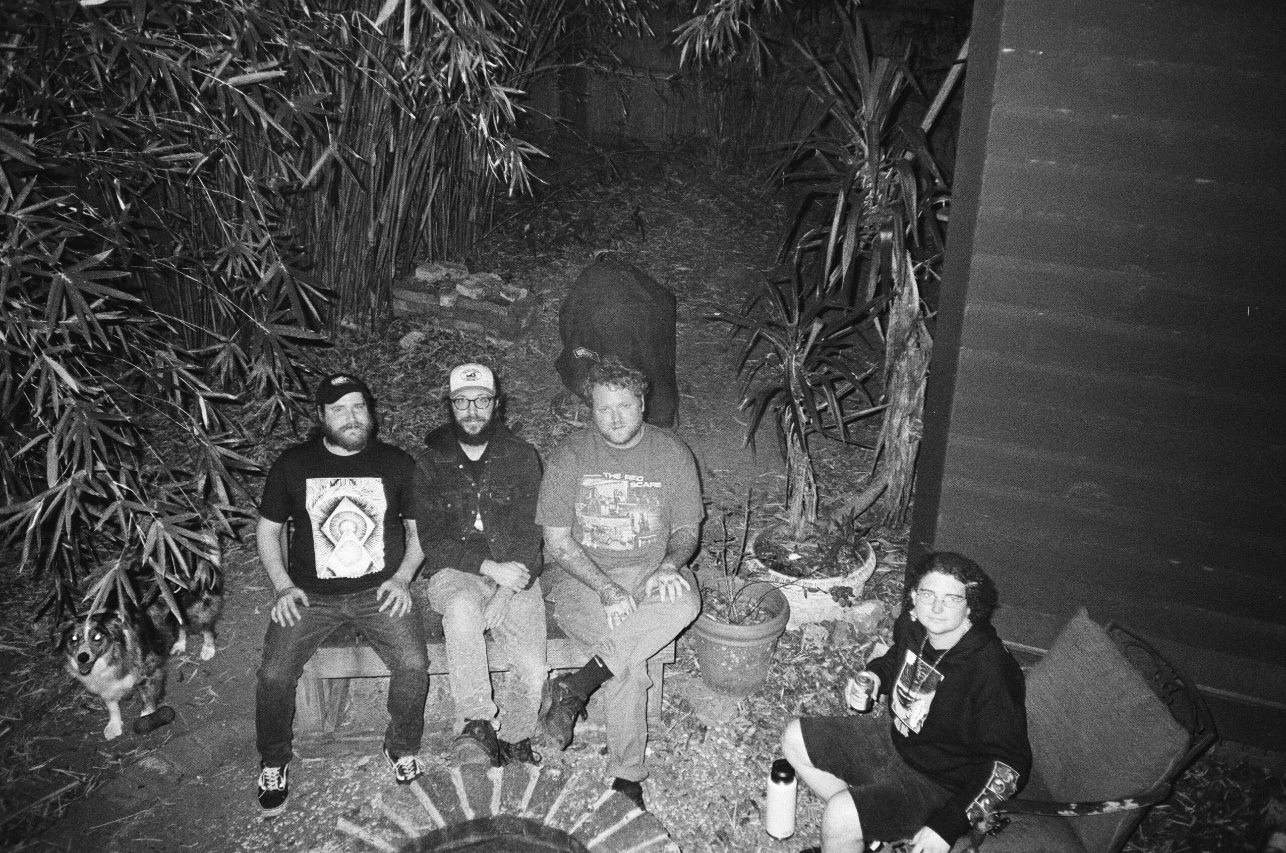Four people sitting outdoors on a circular bench, surrounded by plants and trees, with a person standing behind them.
