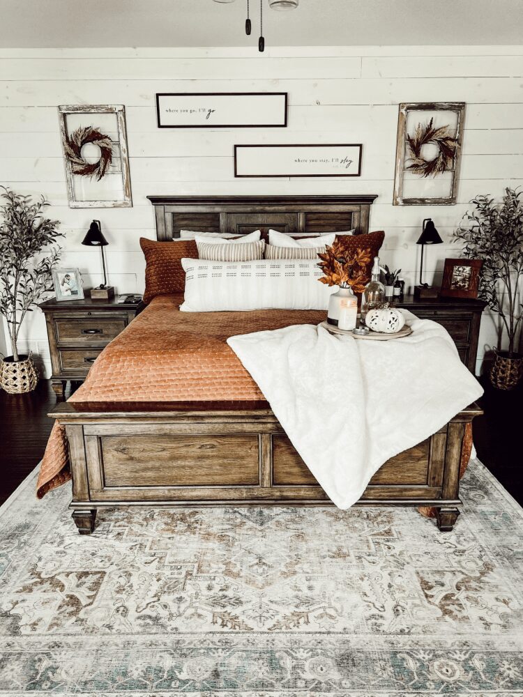 A bedroom with a wooden bed, brown and white pillows, a white blanket, and a tray with flowers and a cup on the bed. Two nightstands with lamps and plants are on either side. Wall art and a white wall are in the background.
