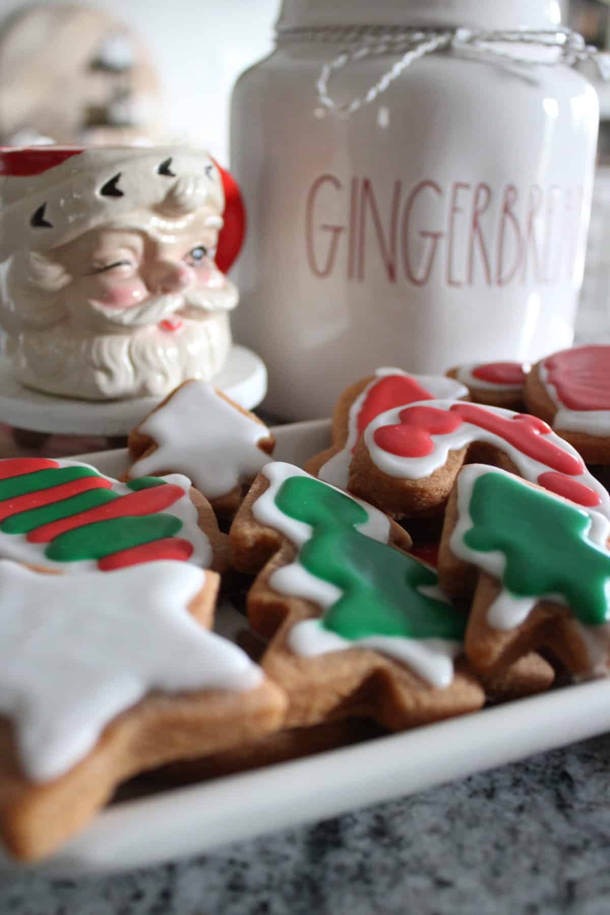 Christmas cookies decorated with green and red icing, a jar labeled 'Gingerbread,' and a Santa figurine, on a gray surface.