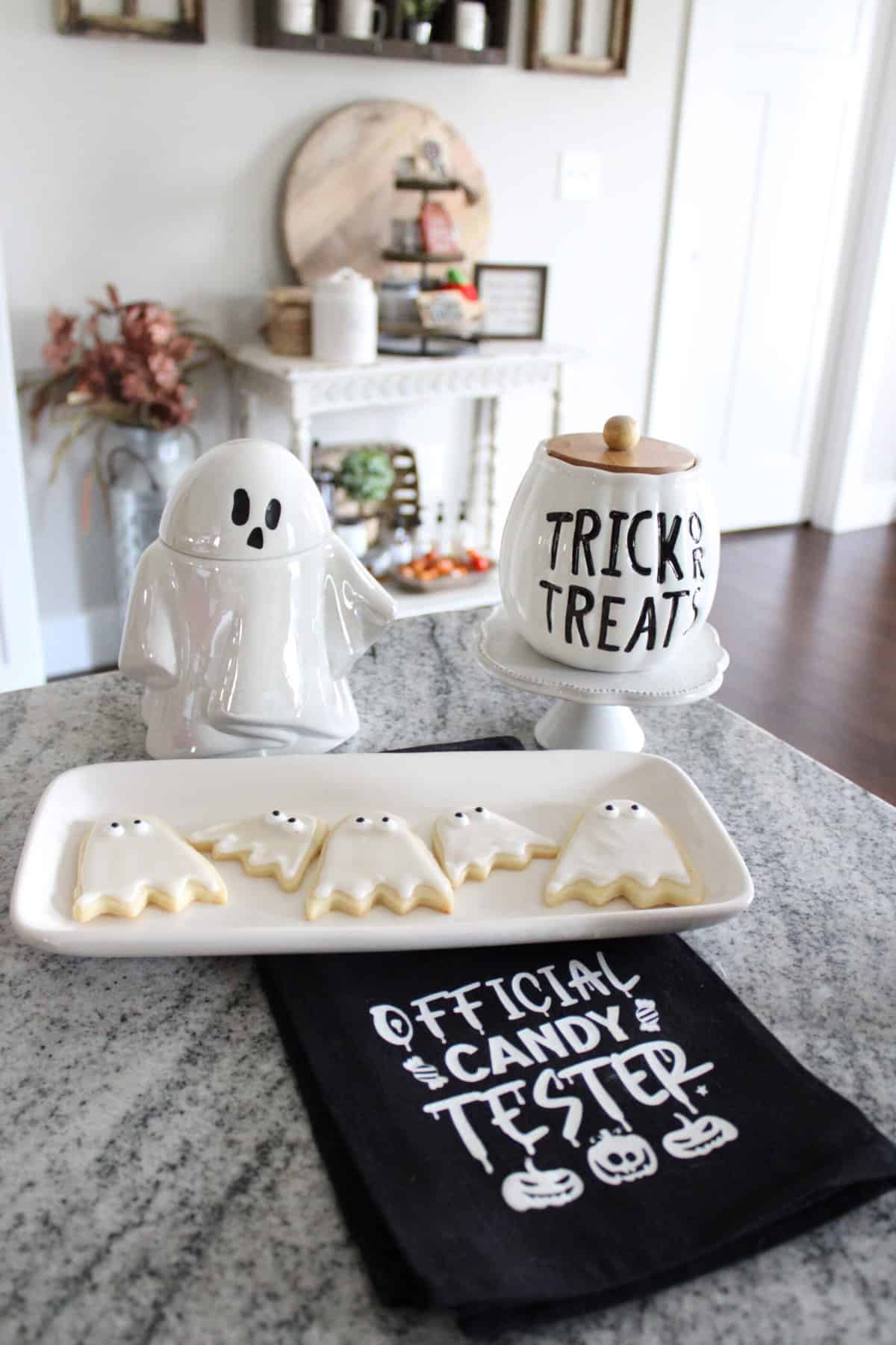 Halloween-themed cookies on a tray, ghost-shaped container, and a jar labeled 'Trick or Treat' on a kitchen counter.