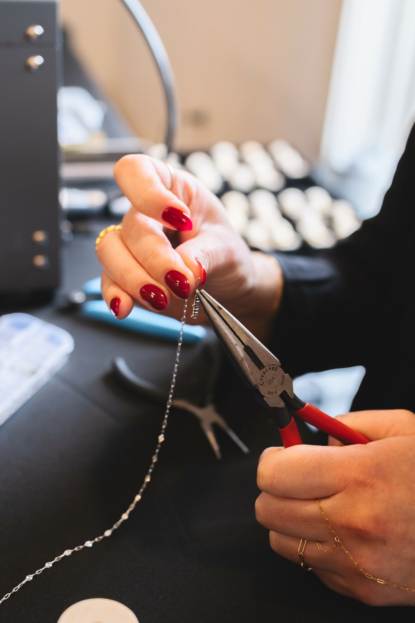 Person's hand with red nail polish cutting a delicate chain with pliers, workspace with jewelry tools in background