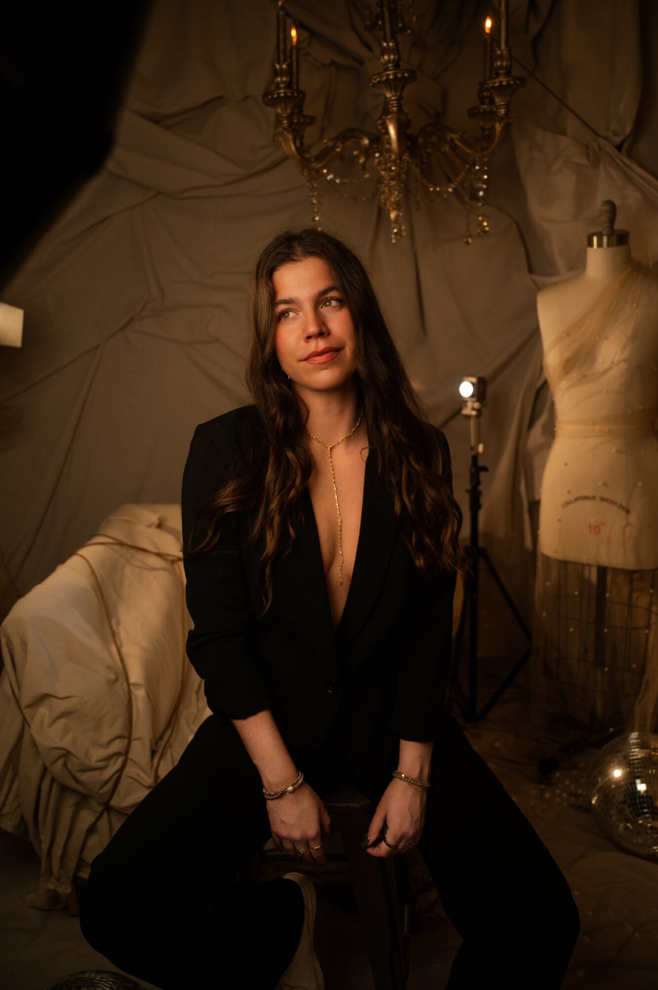 Woman with long dark hair sitting in a dimly lit room, surrounded by vintage decor and a mannequin torso.