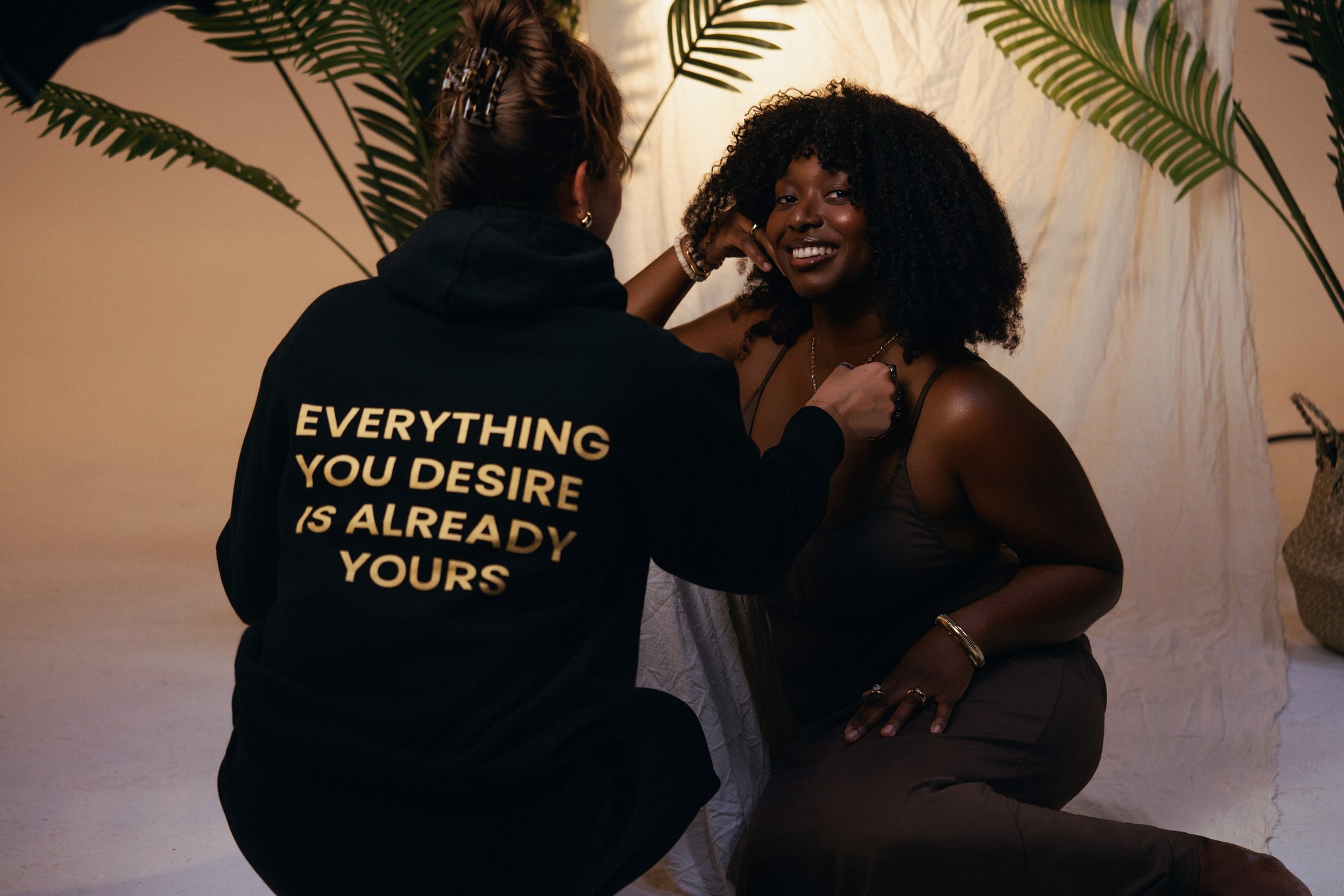 Two women smiling and interacting indoors with plants in the background, one wearing a black hoodie with yellow text.