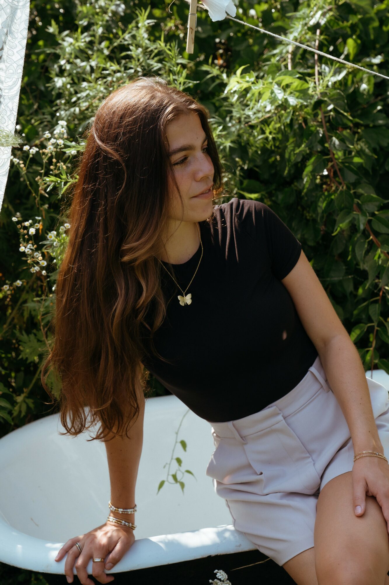 Young woman with long hair wearing a black top and white shorts, standing outdoors among greenery.