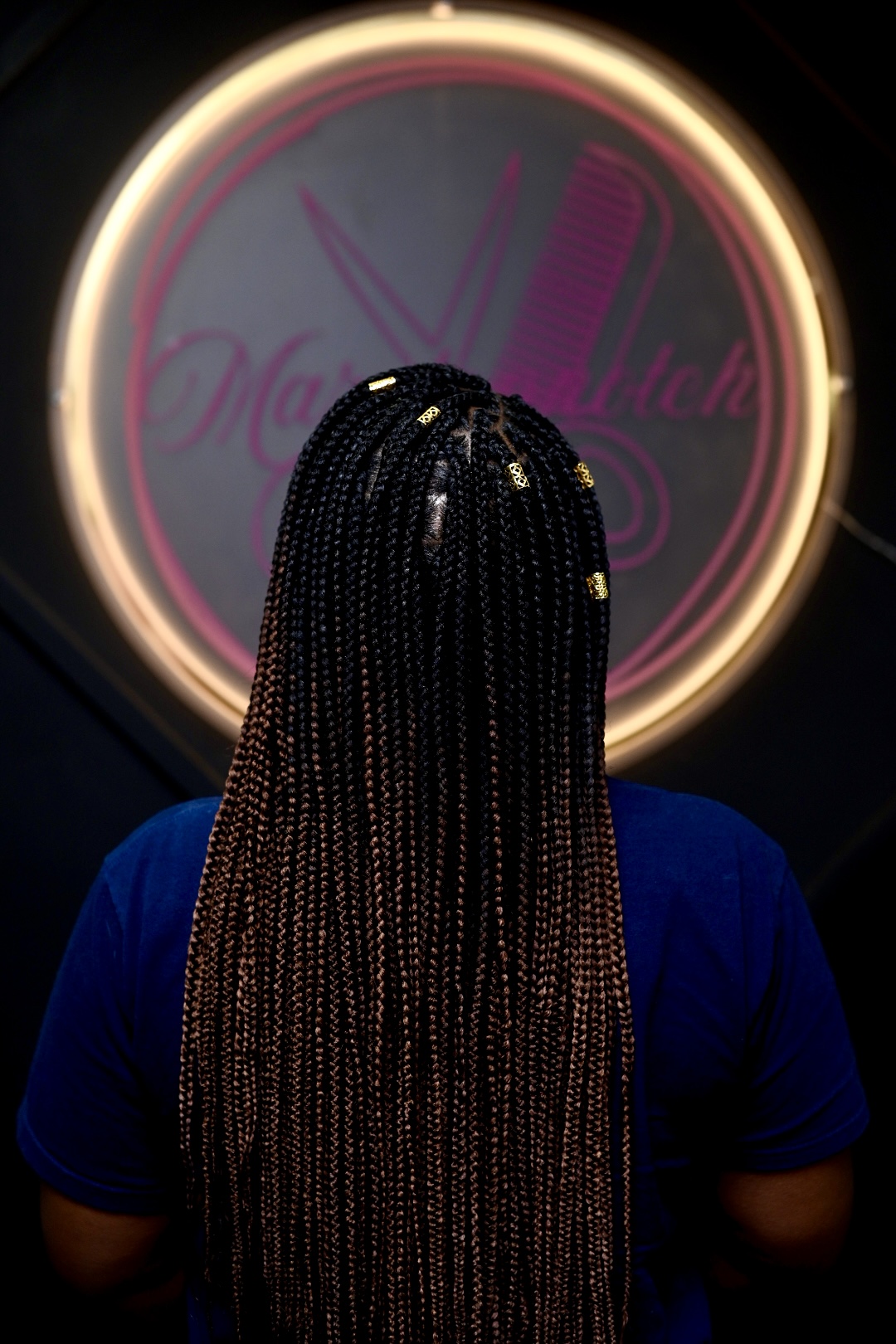 Person with long braided hair facing a neon-lit circular sign, back view, dark background.