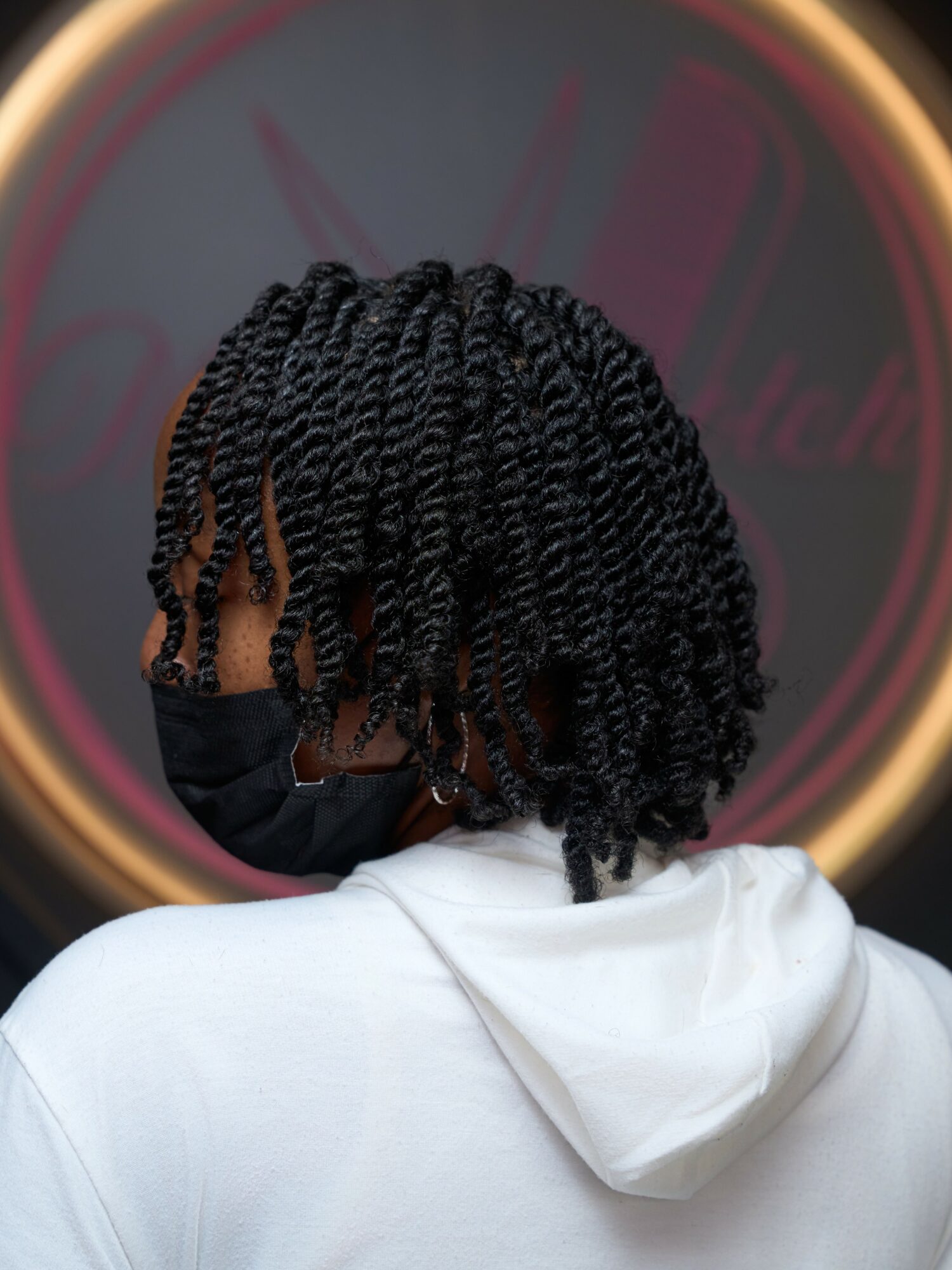 Back view of person with short, twisted hairstyle wearing a face mask and white hoodie, standing in front of a circular sign.