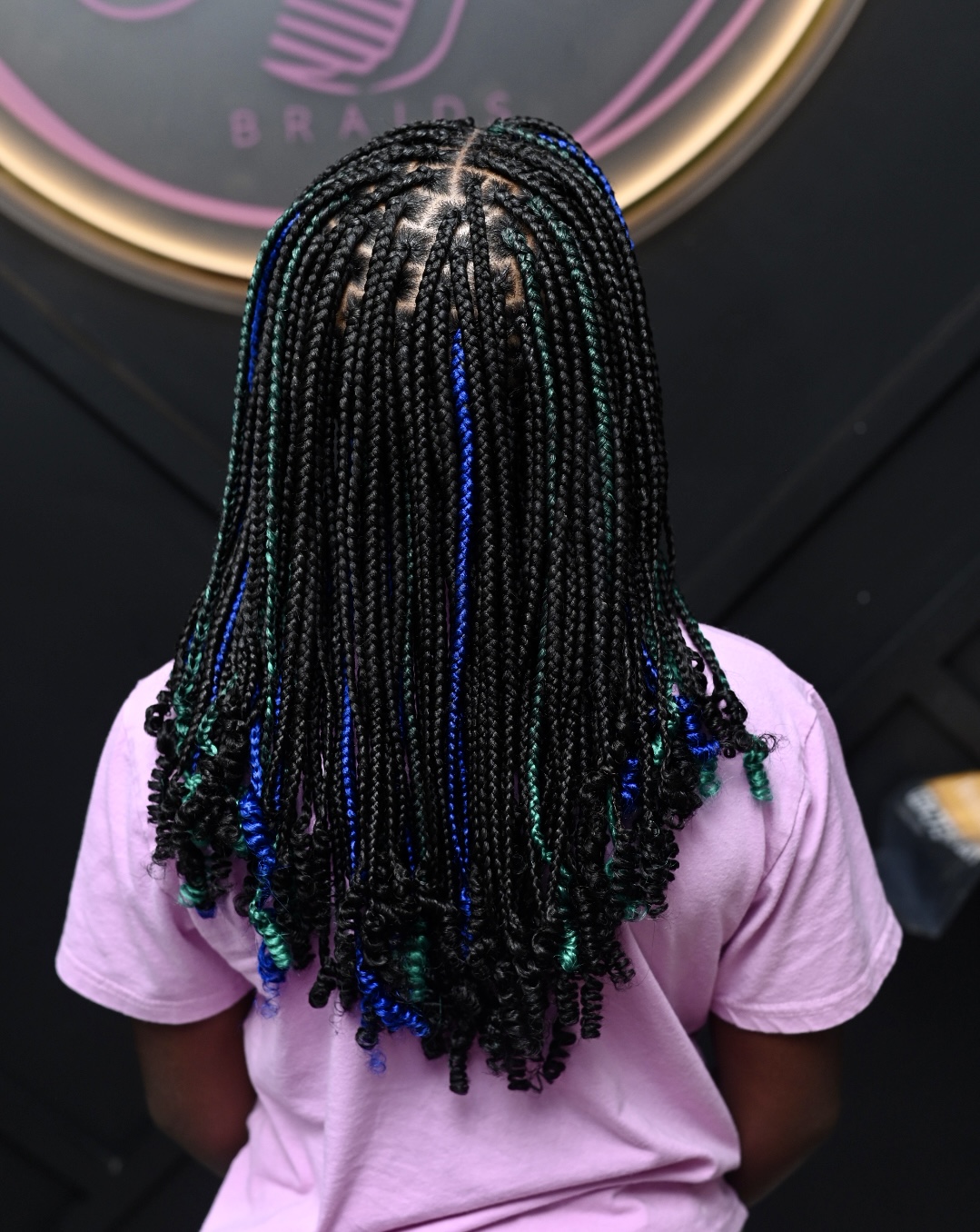 Person with braided hair featuring blue and green accents, wearing a pink shirt, facing away from the camera.