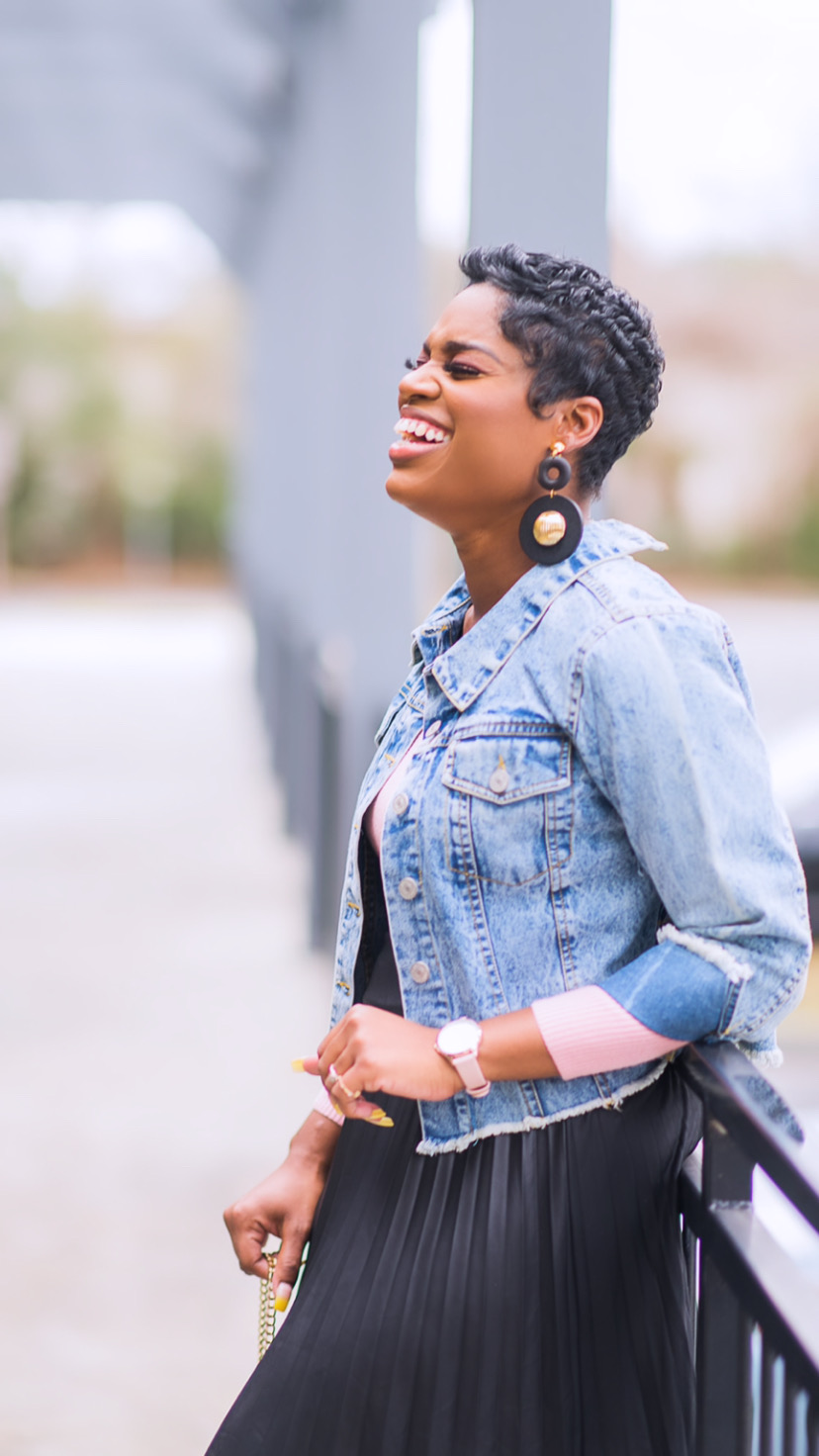Woman with short curly hair smiling, wearing a denim jacket and large earrings, standing outdoors near a railing.
