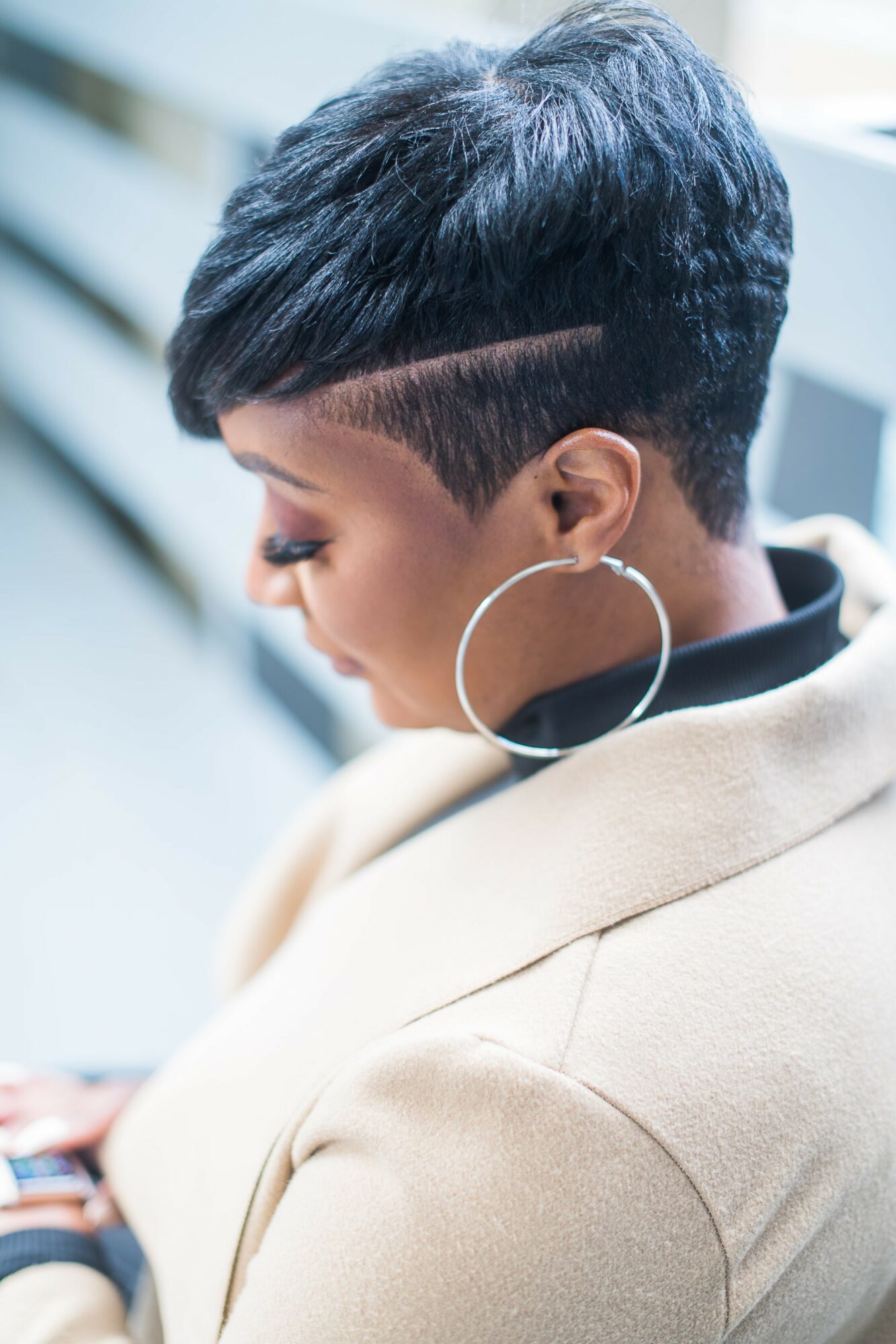 Side view of a woman with a short, textured hairstyle and large hoop earring, wearing a beige coat.