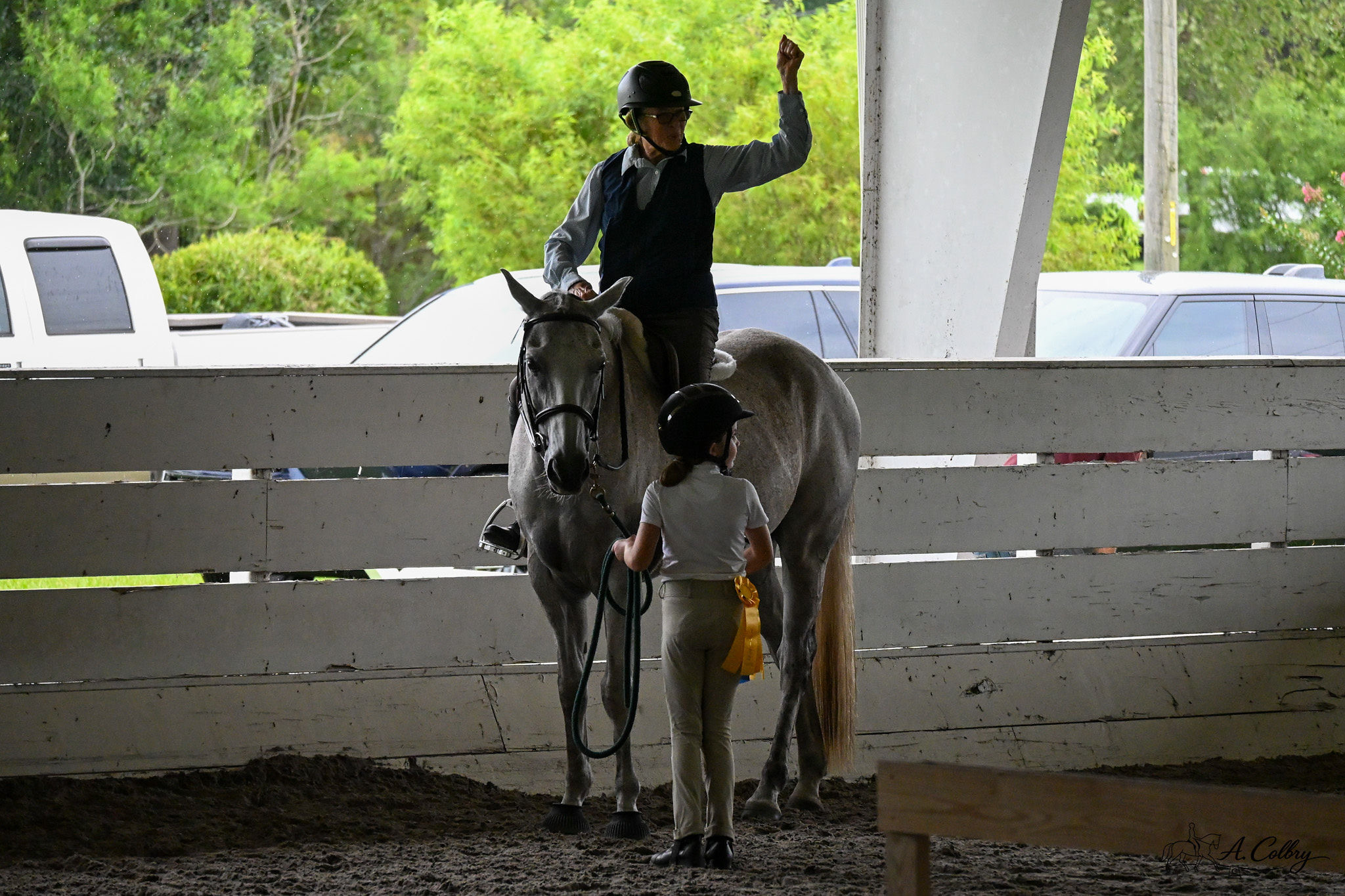 Person riding a horse inside an arena, waving, with a child standing nearby, and a white fence in the background.