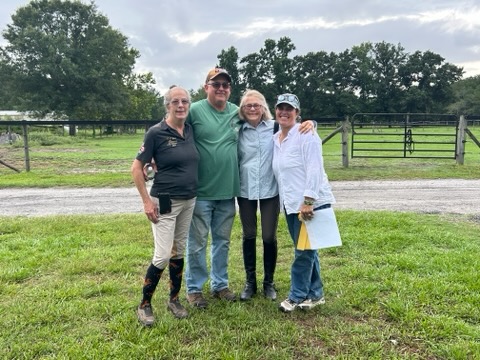 Four people standing outdoors on grass, with trees and a fence in the background, smiling and posing for a photo.