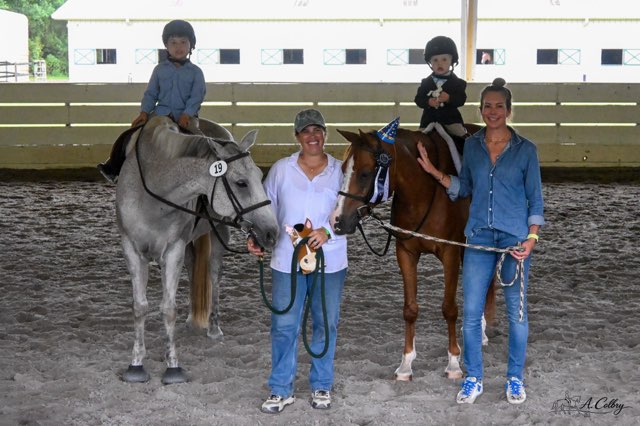 Two children sit on horses, with two women standing beside them holding the horses' reins, in an indoor riding arena.