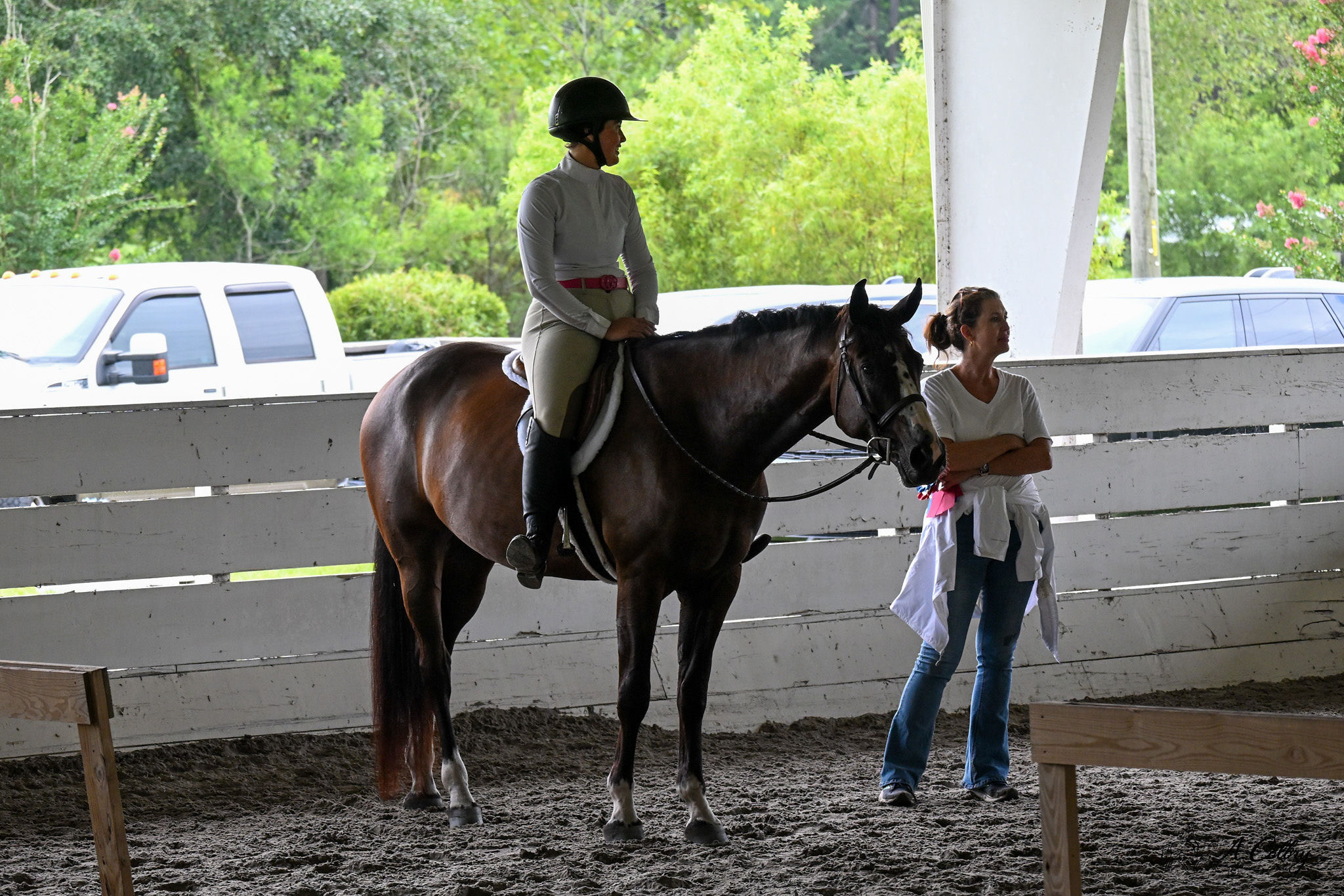Person riding a horse inside an indoor riding arena, with another person standing nearby holding the horse's reins, and vehicles outside visible through windows.