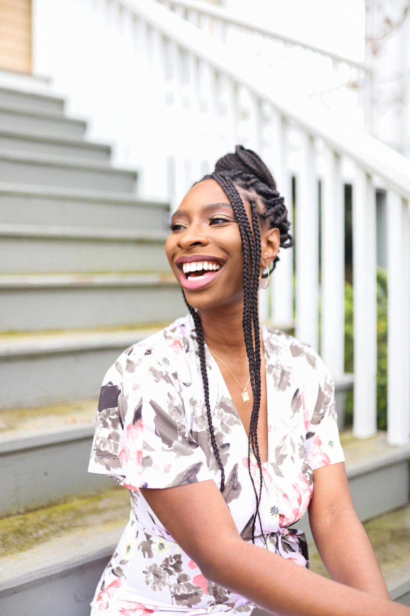 Smiling woman with braided hair sitting on outdoor stairs, wearing a floral shirt, white railing in background.