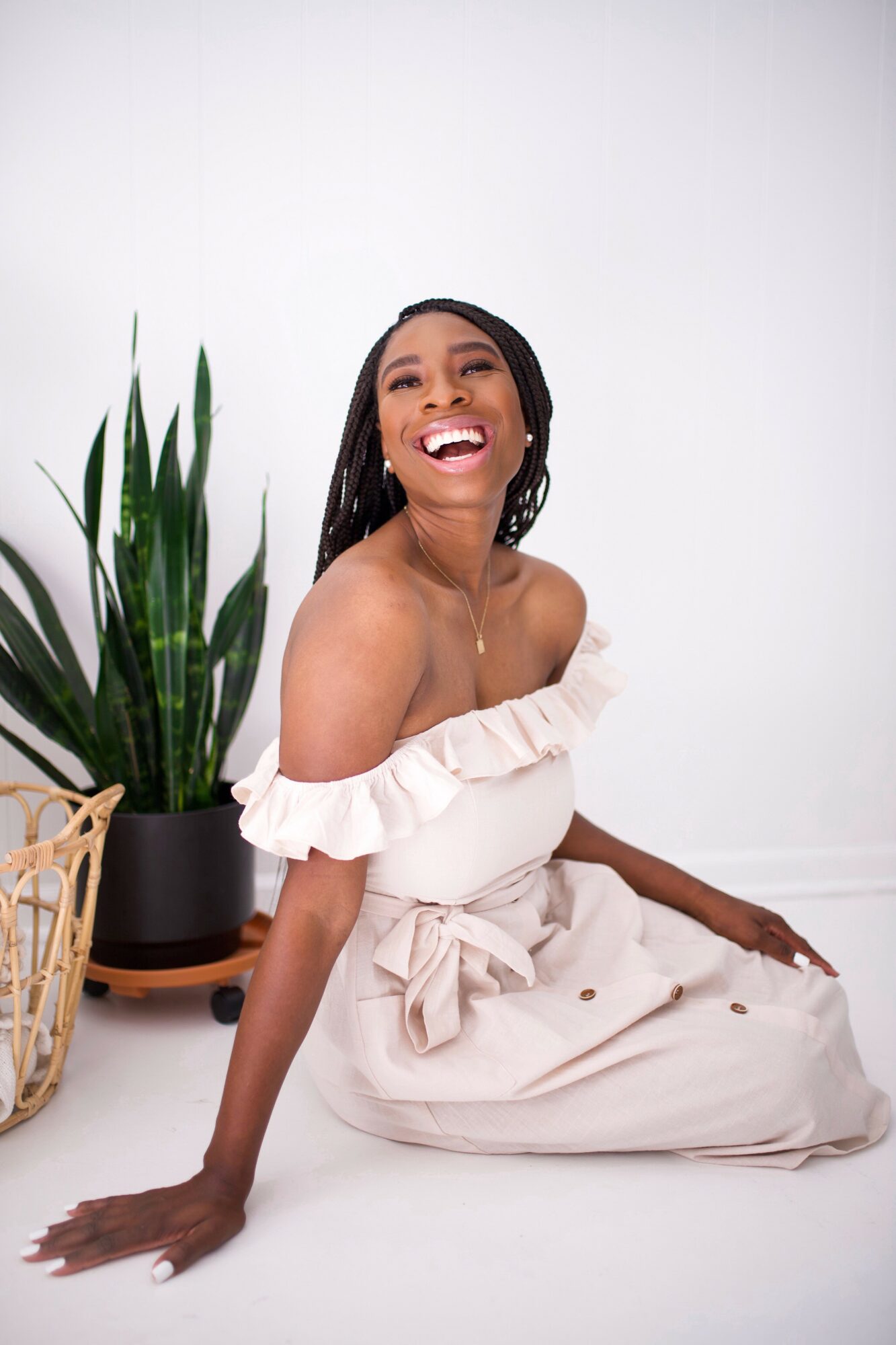Smiling woman sitting on the floor next to a potted plant, wearing an off-shoulder dress with ruffles.