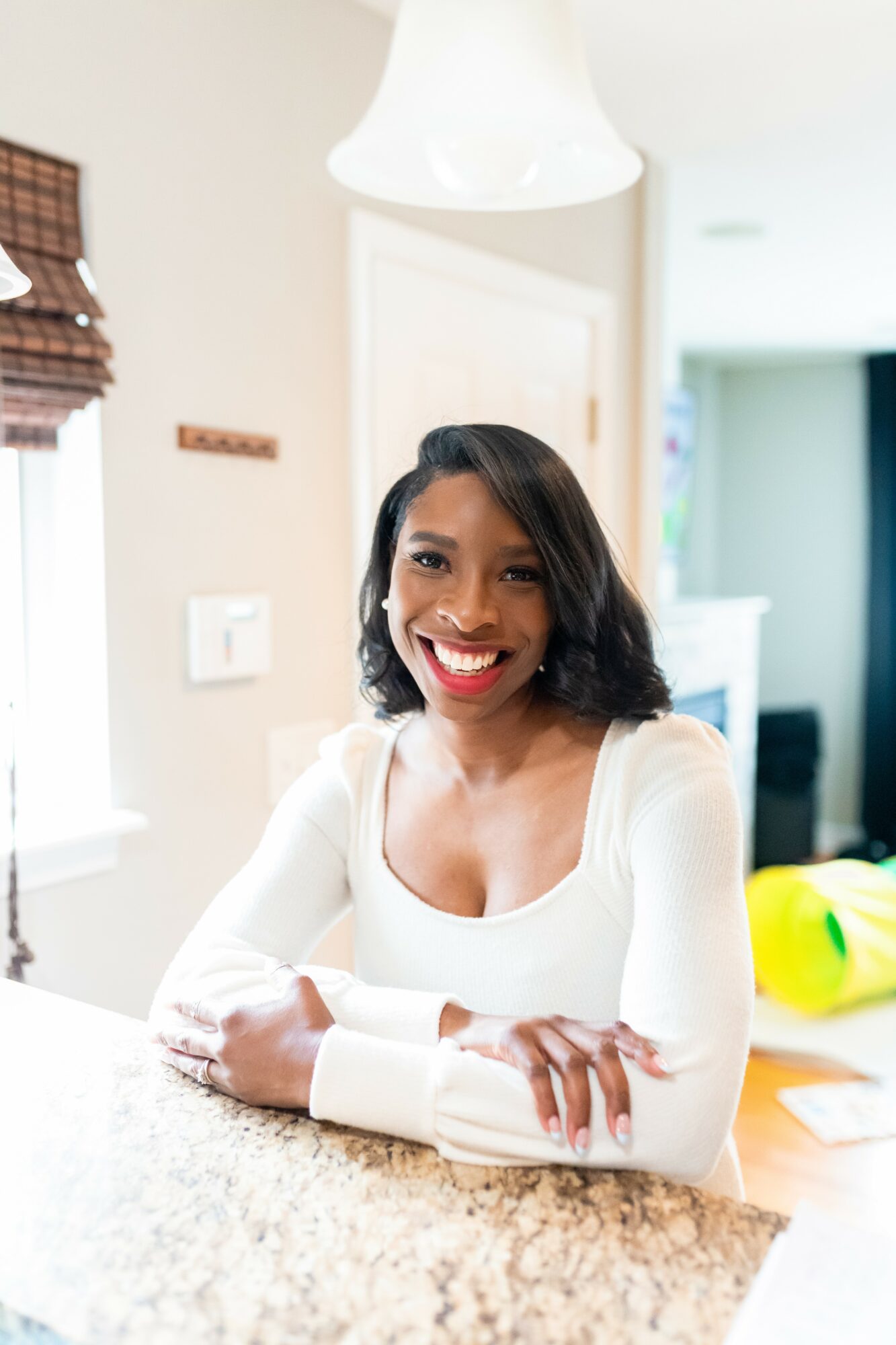 Smiling woman with shoulder-length dark hair, wearing a white top, sitting at a kitchen counter with a bright background.