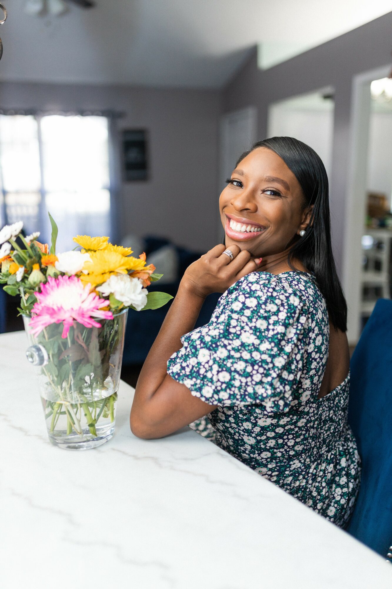 Smiling woman with long dark hair sitting at a table with a colorful flower arrangement, in a bright room.