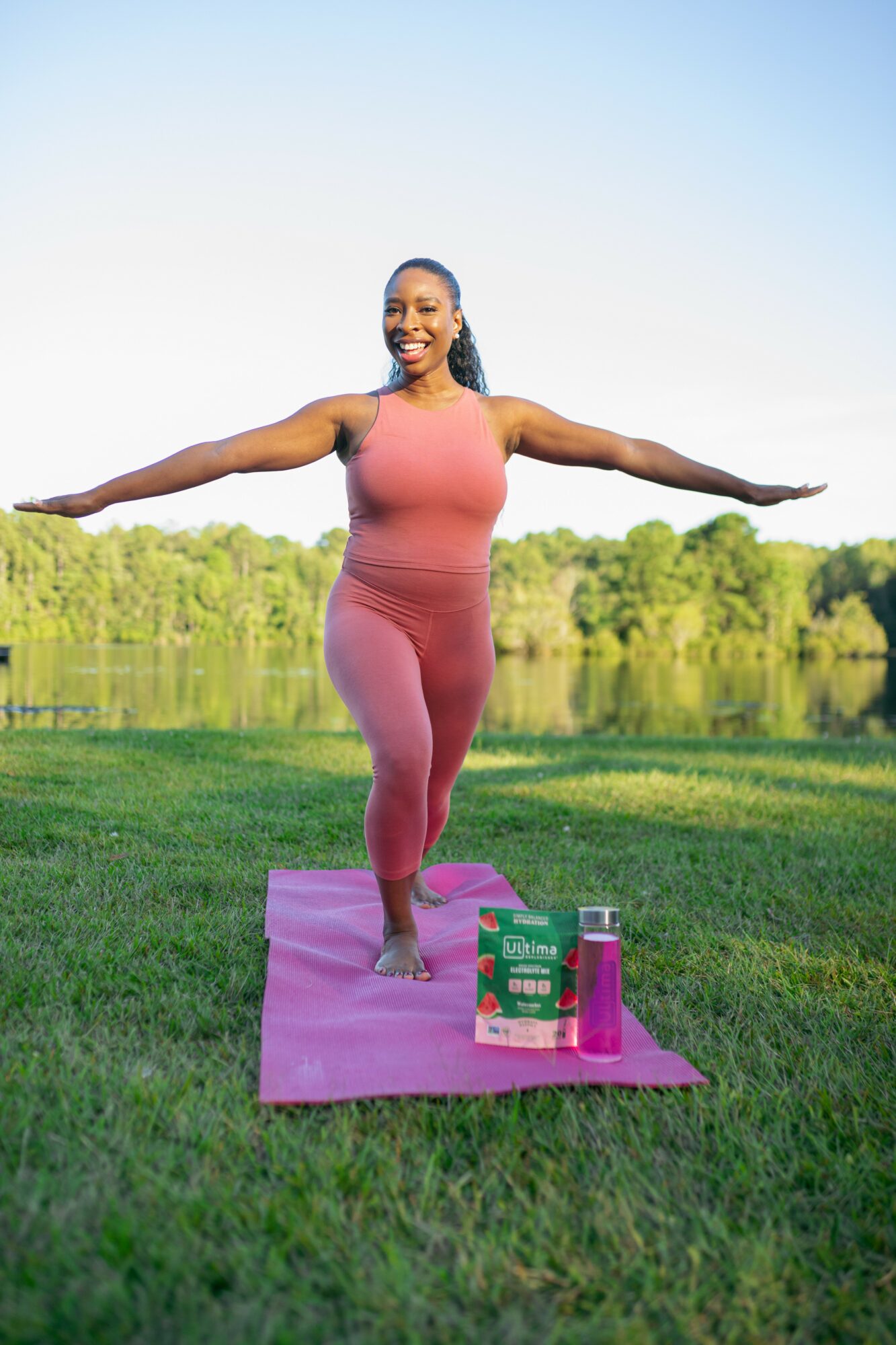 Woman practicing yoga outdoors on a pink mat, arms extended, smiling, with a lake and trees in the background.