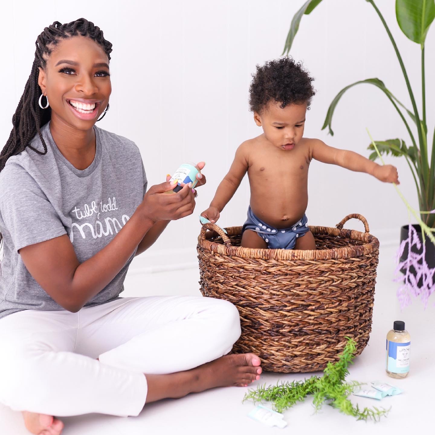 Woman smiling with a child in a basket, holding a bottle, surrounded by plants and bottles, indoors.