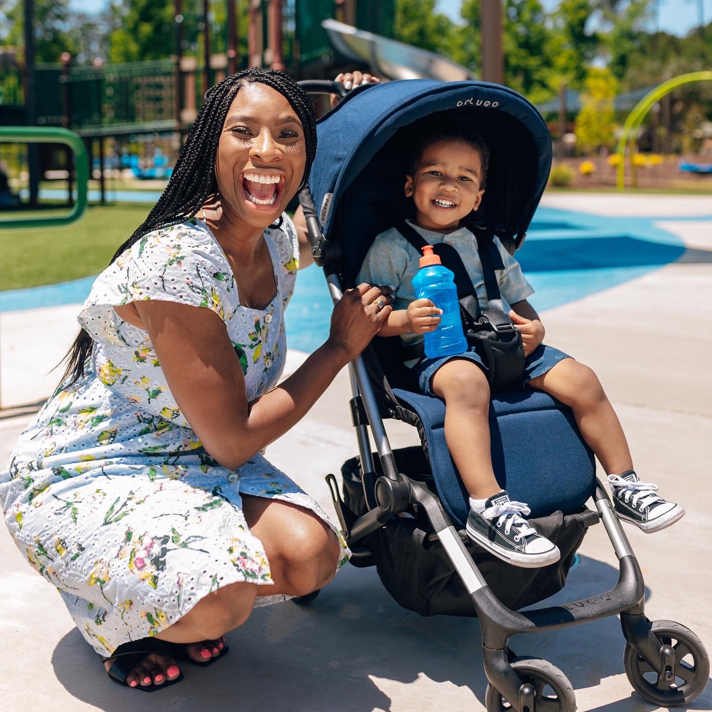 Woman crouching next to a smiling child in a stroller at a park, holding a water bottle, sunny day, trees in background.