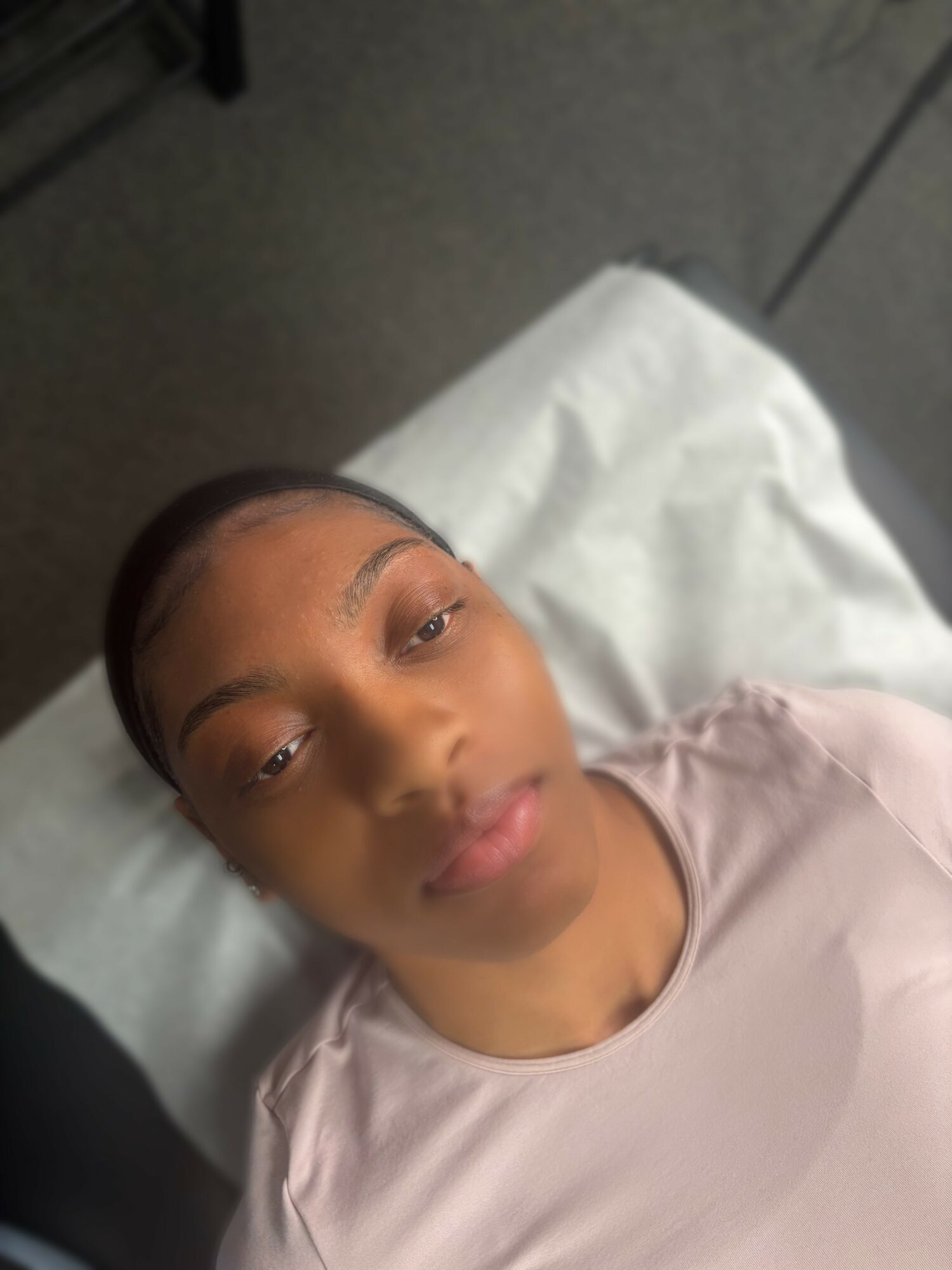 Young person lying on a bed with a white pillow, looking at the camera, wearing a light-colored shirt.