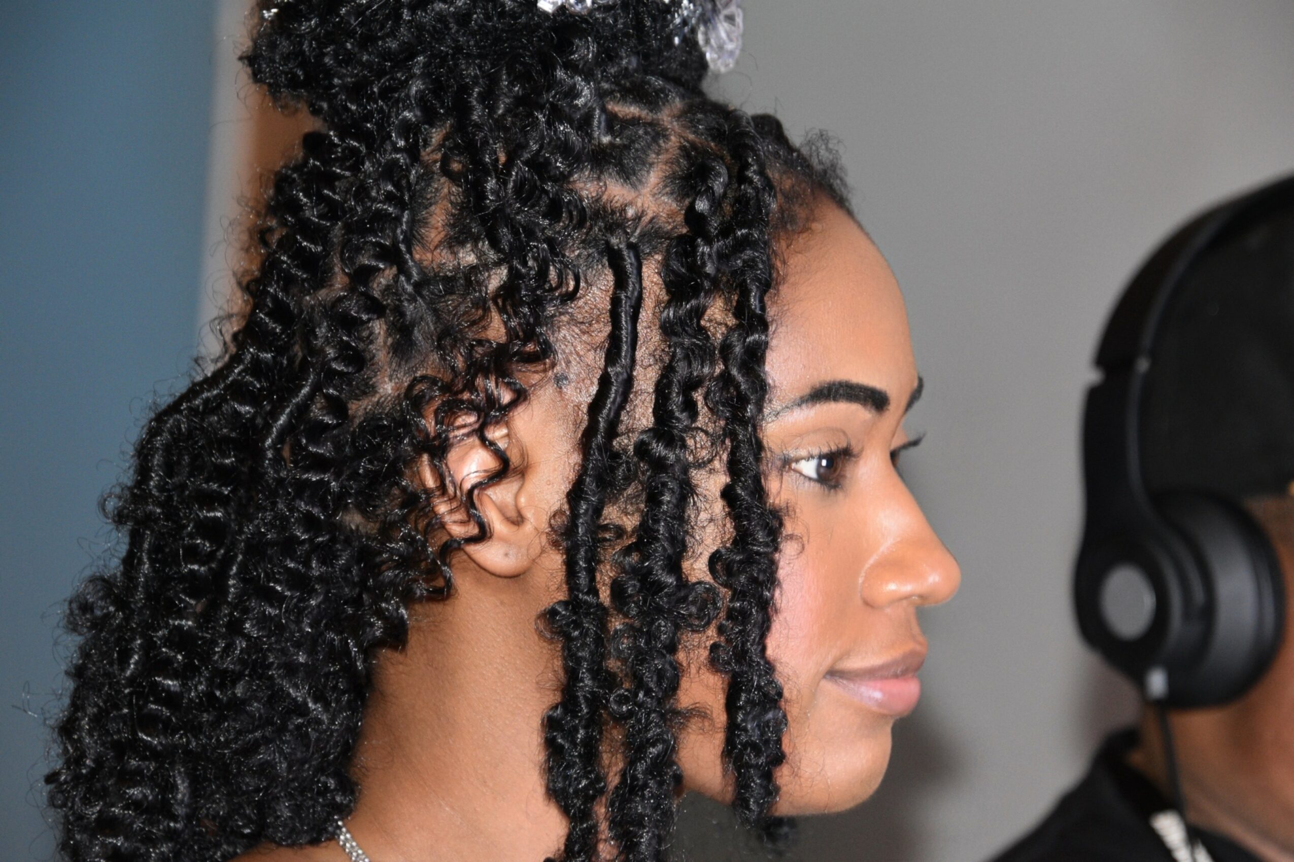 Close-up of a woman with curly hair looking to the right, with a person wearing headphones partially visible.