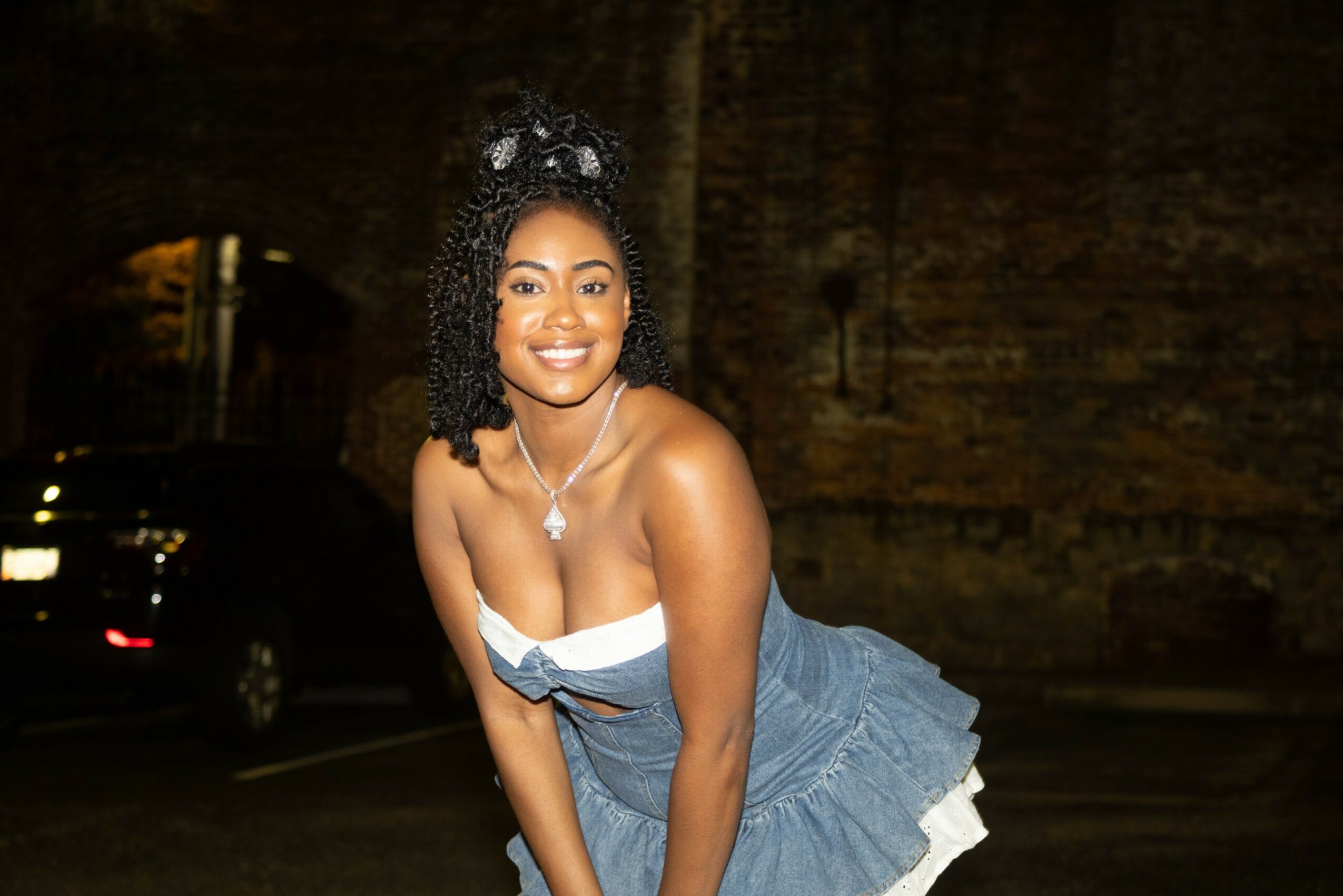 Smiling woman in a strapless blue dress with white accents, standing outdoors at night with a brick wall background.