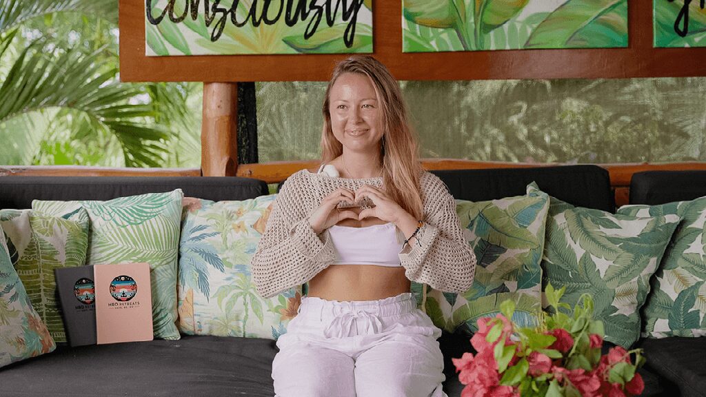 Young woman sitting on a sofa with floral cushions, smiling and making a heart shape with her hands, in a bright room.