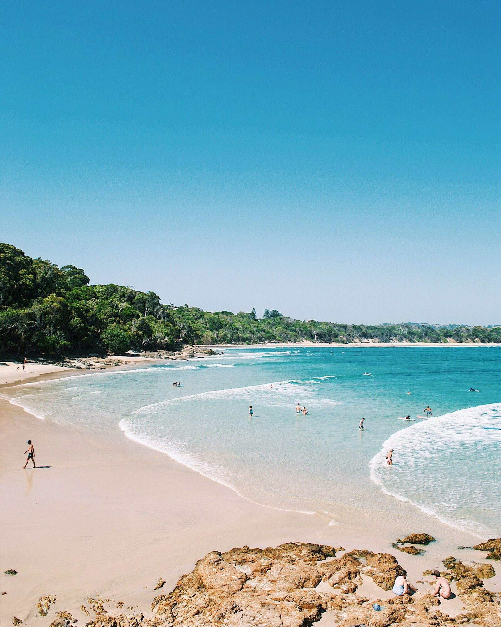 Sunny beach with clear blue sky, sandy shore, and people swimming and walking along the coast.