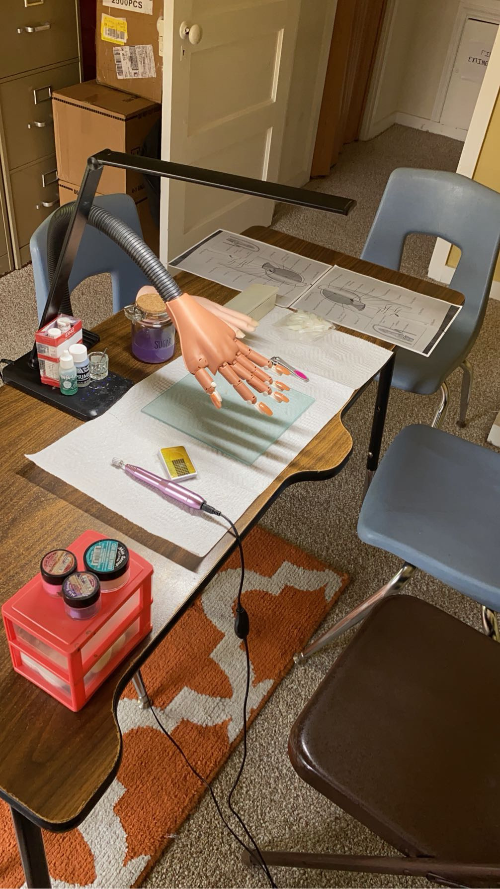 Table with a mannequin hand, papers, and various objects, in a room with chairs and a door, viewed from the side.