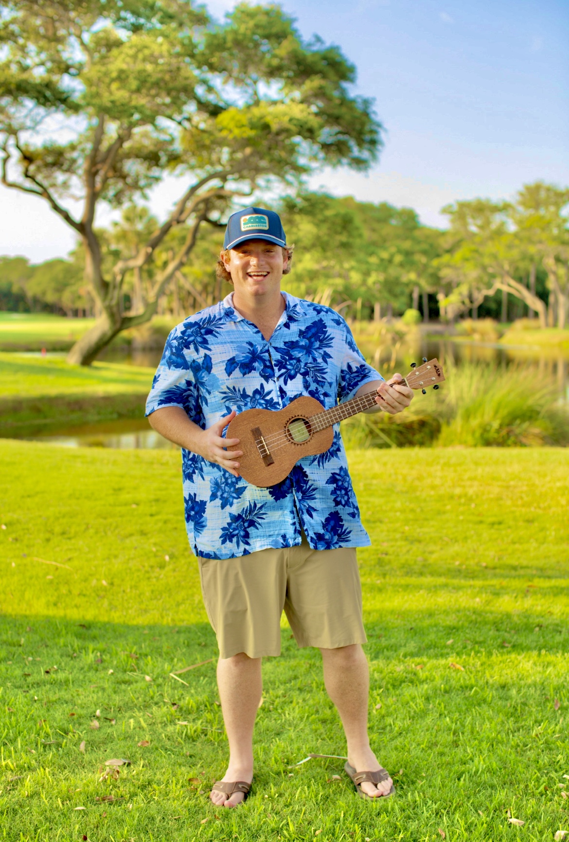 Young man standing on grass holding a ukulele, wearing a blue floral shirt, beige shorts, and a cap, outdoors with trees and water in background.
